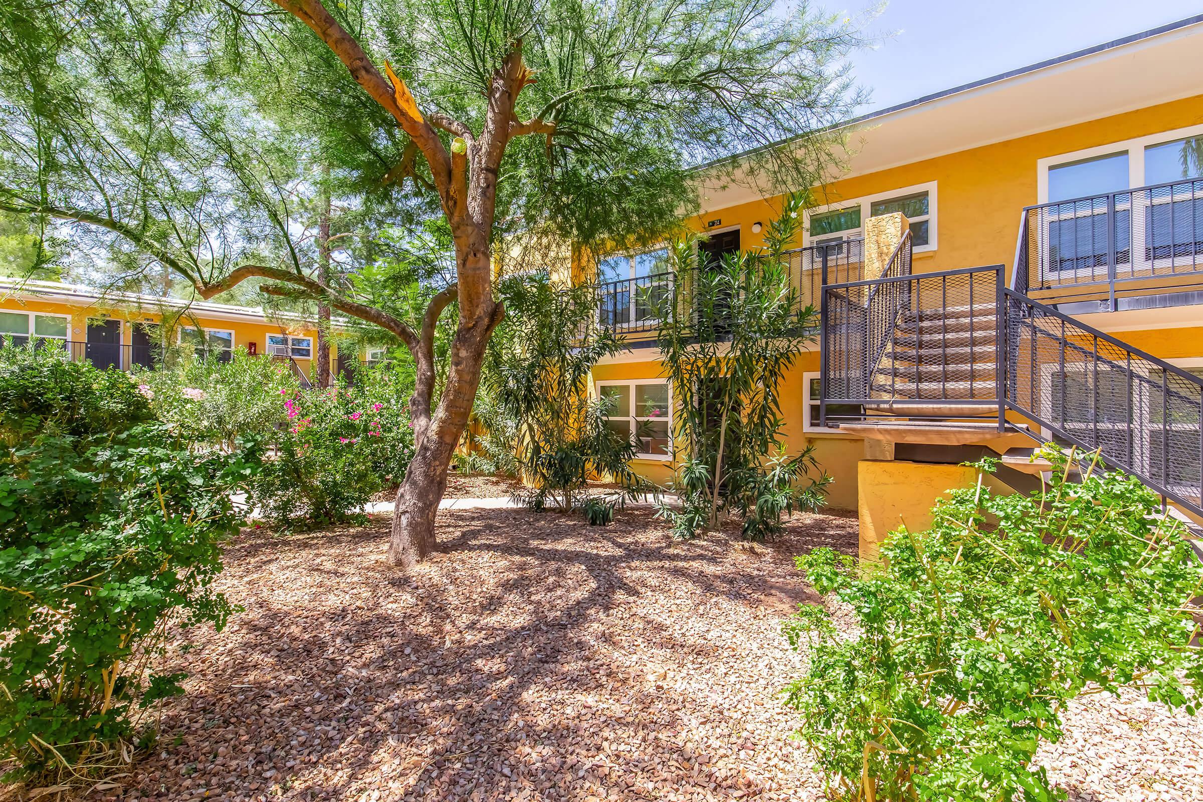 A vibrant courtyard featuring a yellow apartment building surrounded by lush greenery, including small trees and flowering plants. A staircase leads to the upper level of the building, with rocky landscaping creating a natural feel in the outdoor space.