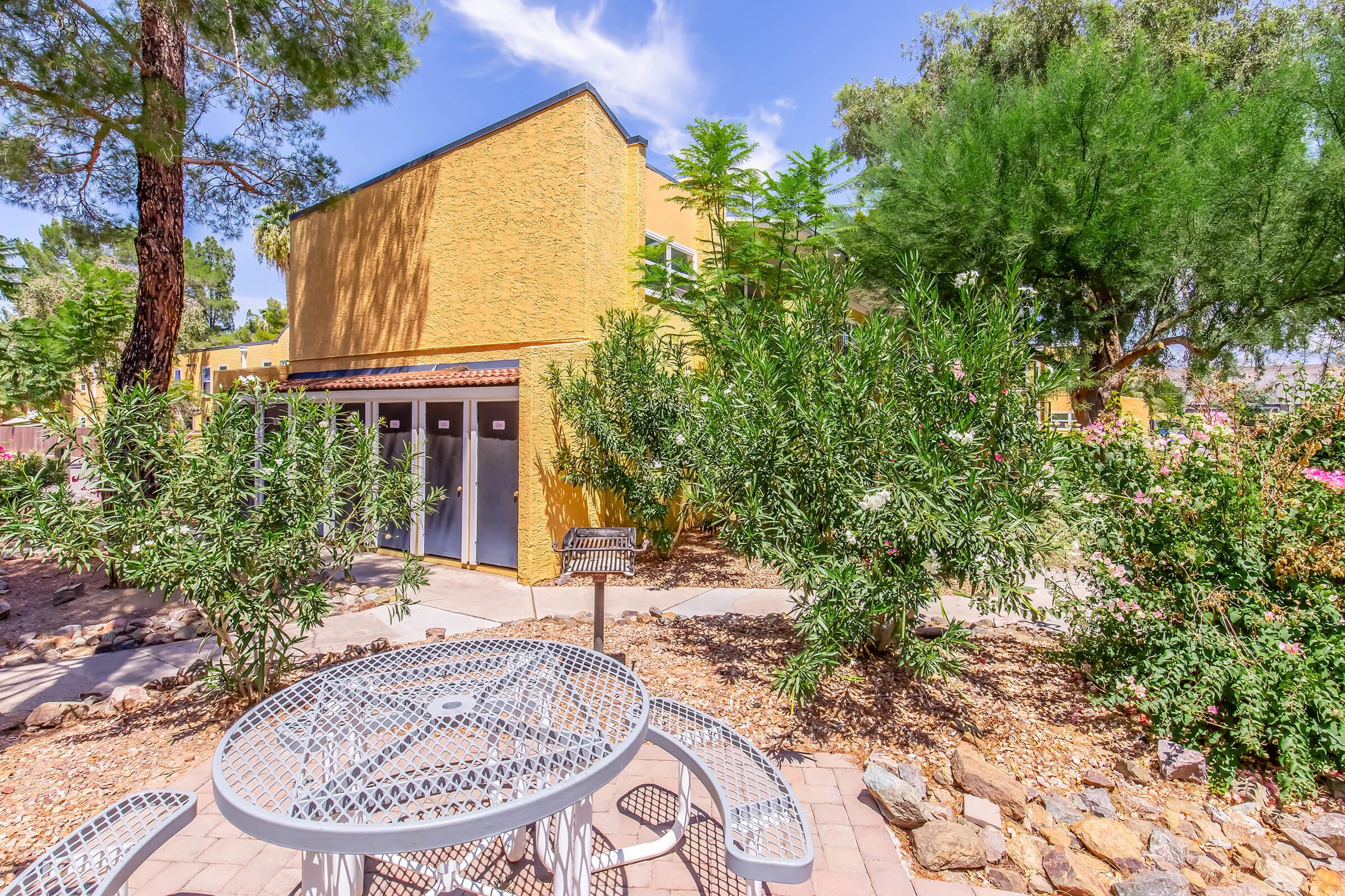 A sunny outdoor area featuring a yellow building surrounded by greenery, including shrubs and trees. In the foreground, there is a round metal table with chairs, and a small barbecue grill nearby. The sky is partly cloudy, adding to the pleasant atmosphere.