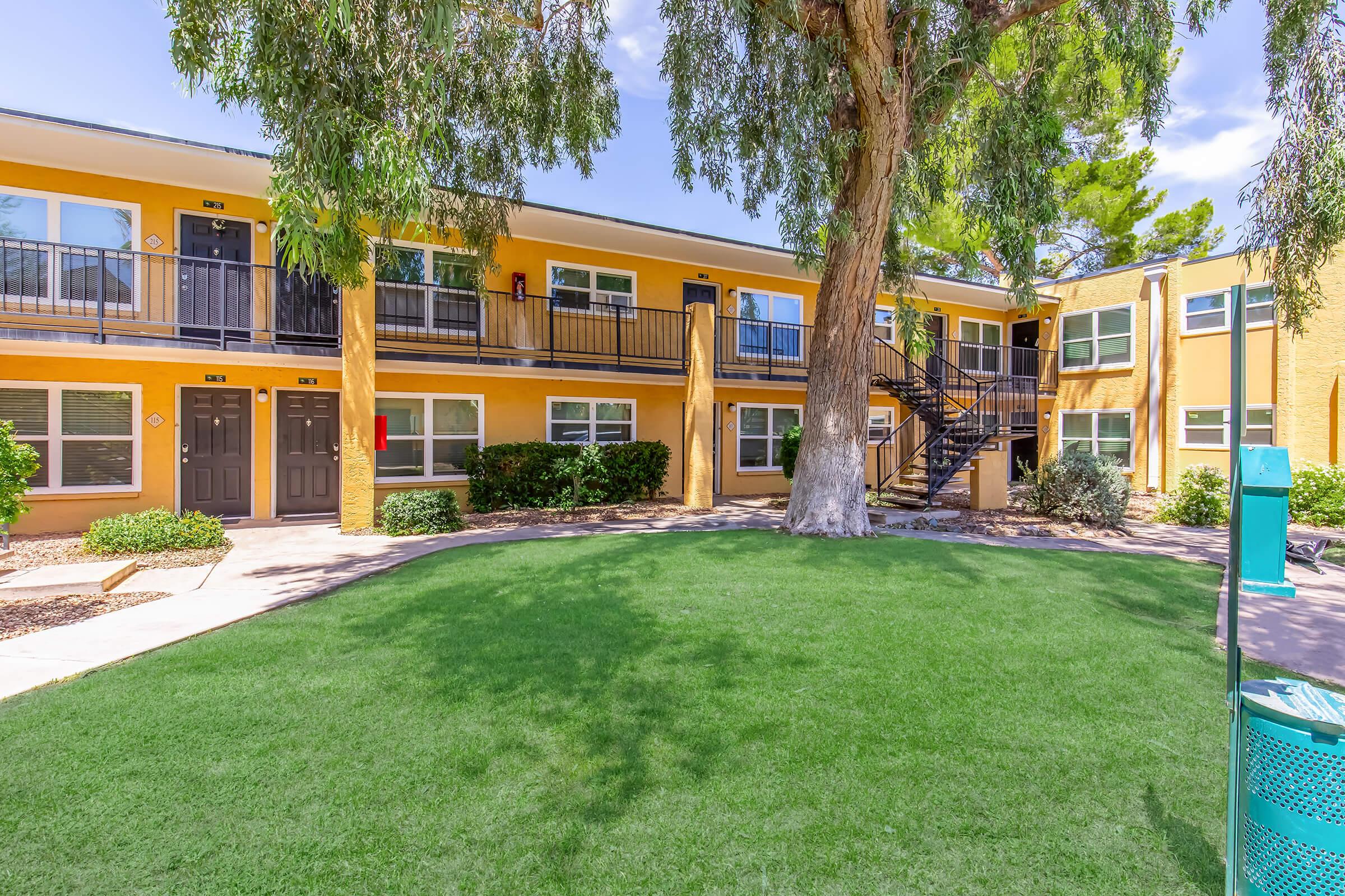 A well-maintained apartment complex with two-story buildings painted in yellow. The scene includes green grass, landscaping around the building, and a set of outdoor stairs leading to the upper level. Nearby, there is a trash can and trees providing shade. Blue sky with few clouds in the background.