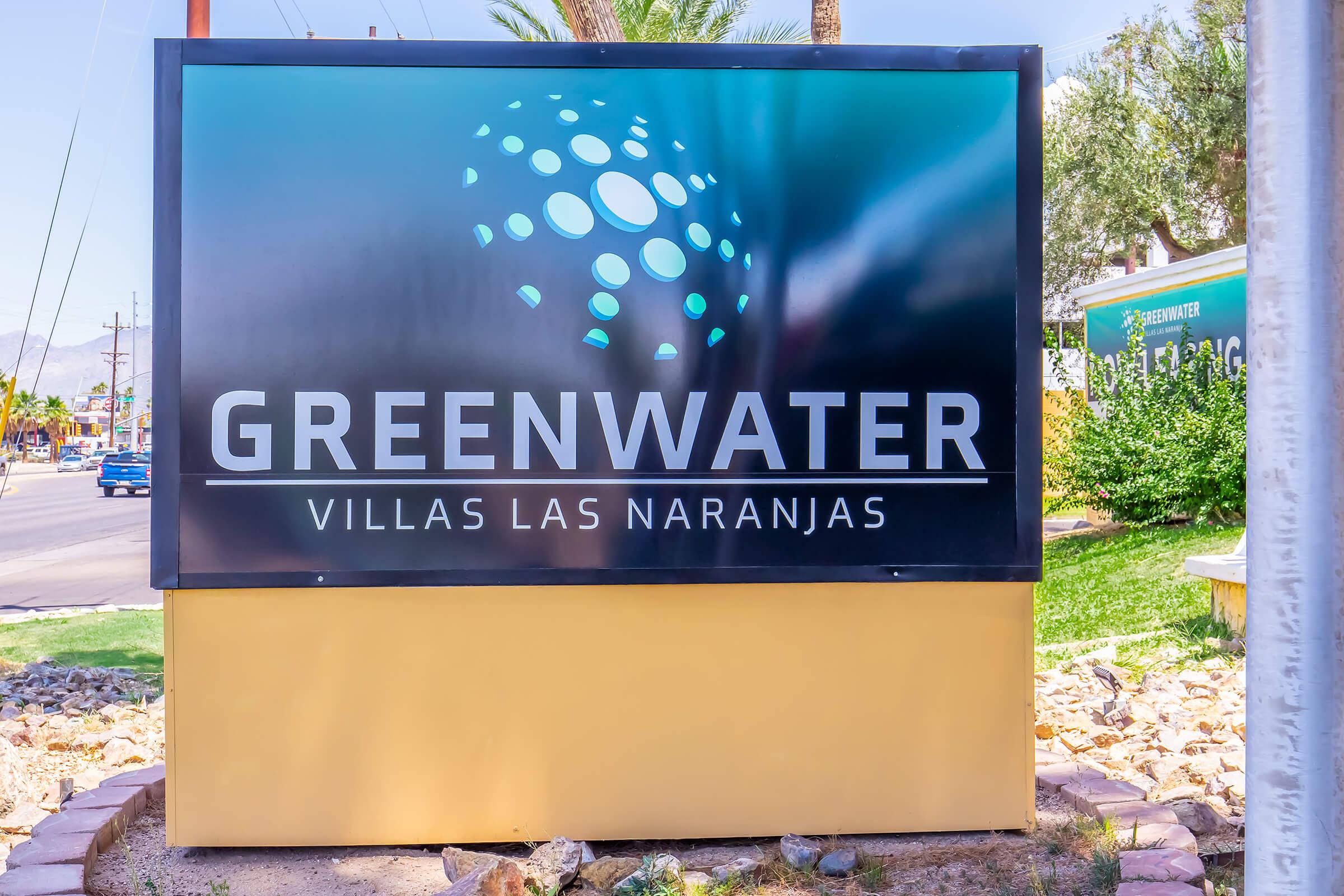 Sign for Greenwater Villas Las Naranjas, featuring a modern design with circular graphics and text. The sign is placed outdoors with palm trees in the background, indicating a residential or vacation property. Bright colors and clear branding are prominent.