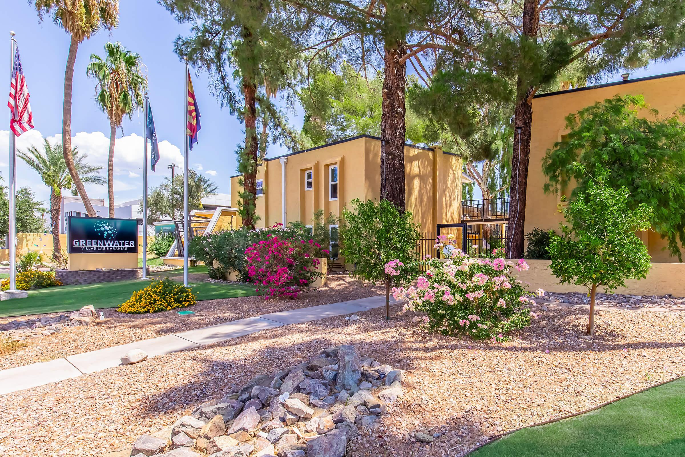 Landscape view of a property entrance featuring colorful flower beds, desert plants, and a sign labeled "GREENWATER." In the background, there are several palm trees and flags, indicating a sunny day in a warm climate. The scene highlights well-maintained surroundings and a welcoming atmosphere.