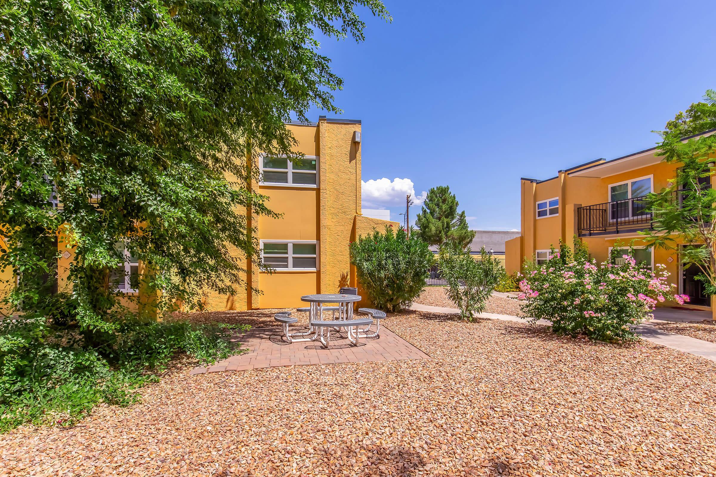 A peaceful outdoor area featuring a small round table with benches surrounded by landscaped pathways and decorative gravel. Lush green bushes and flowering plants add color, while two yellow buildings with large windows are visible in the background under a clear blue sky.