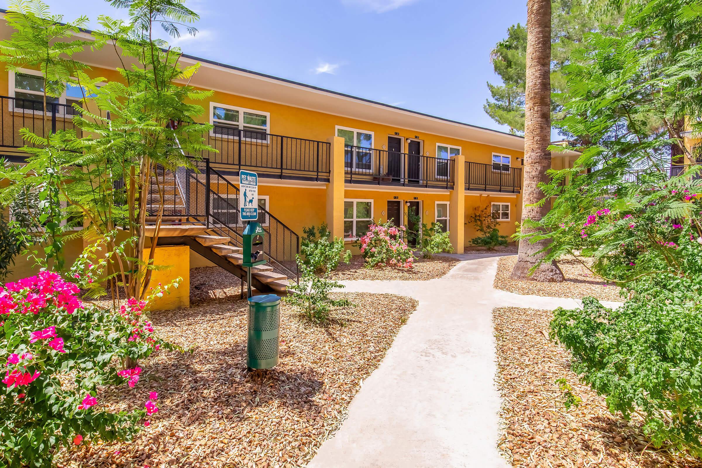 A colorful two-story apartment building with a bright yellow exterior, surrounded by blooming flowers and greenery. A pathway leads through the landscaping, and there are stairs leading to the upper level. Palm trees are in the background, contributing to a tropical ambiance.