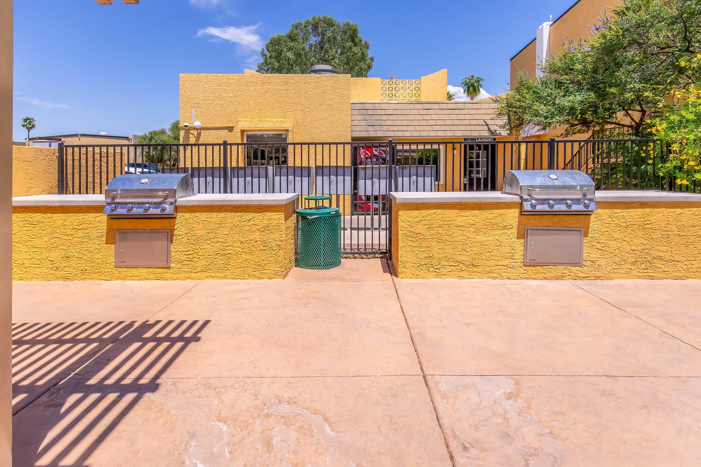 Outdoor grilling area featuring two stainless steel gas grills on a stone countertop. The backdrop includes a bright yellow wall and a glimpse of a building with greenery and a blue sky. A green trash bin is positioned next to the grills on the paved surface.