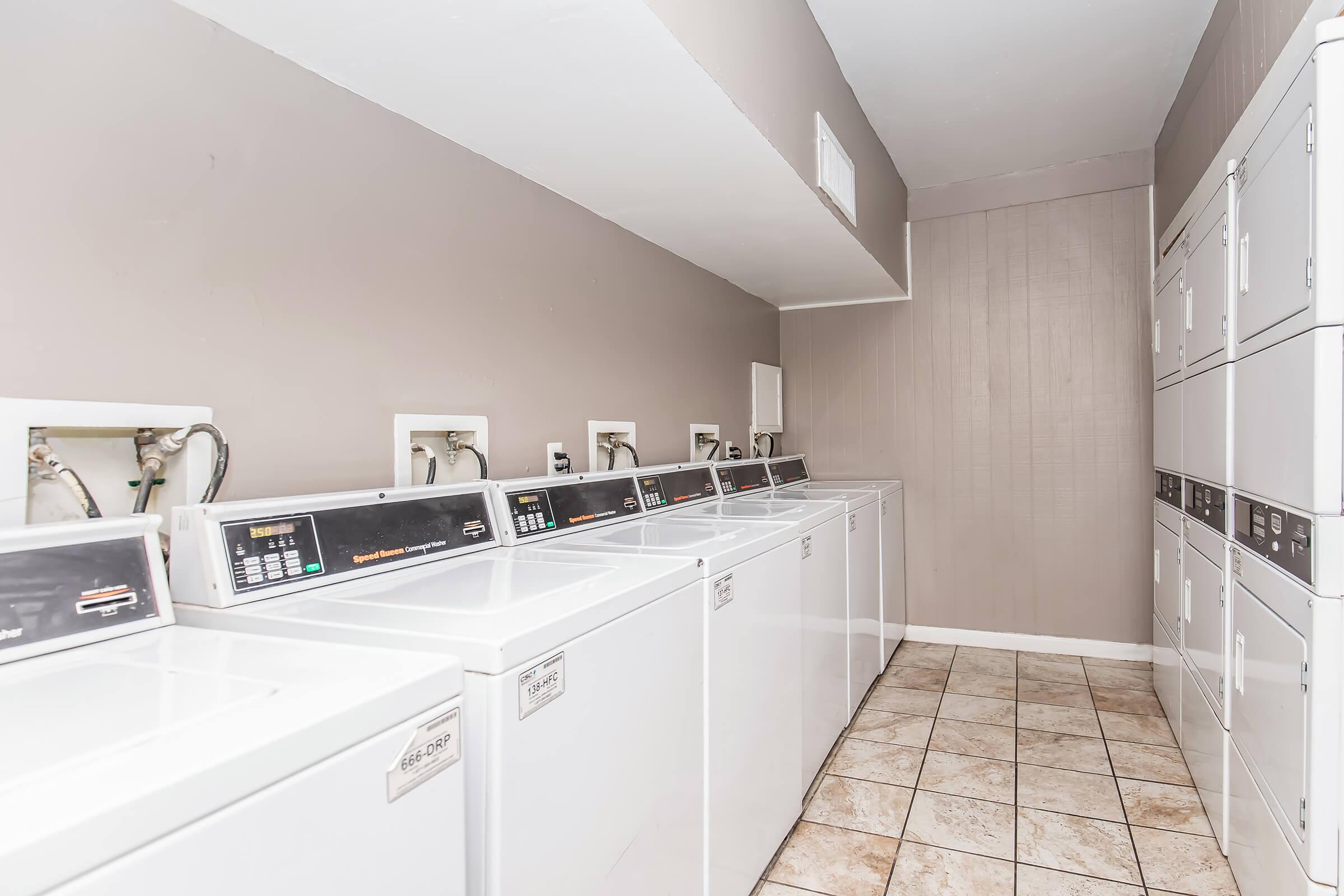 A clean, well-lit laundry room featuring several rows of white washing machines and stacked dryers. The floor is tiled, and the walls are painted in a neutral color, creating a spacious and organized environment for laundry tasks.