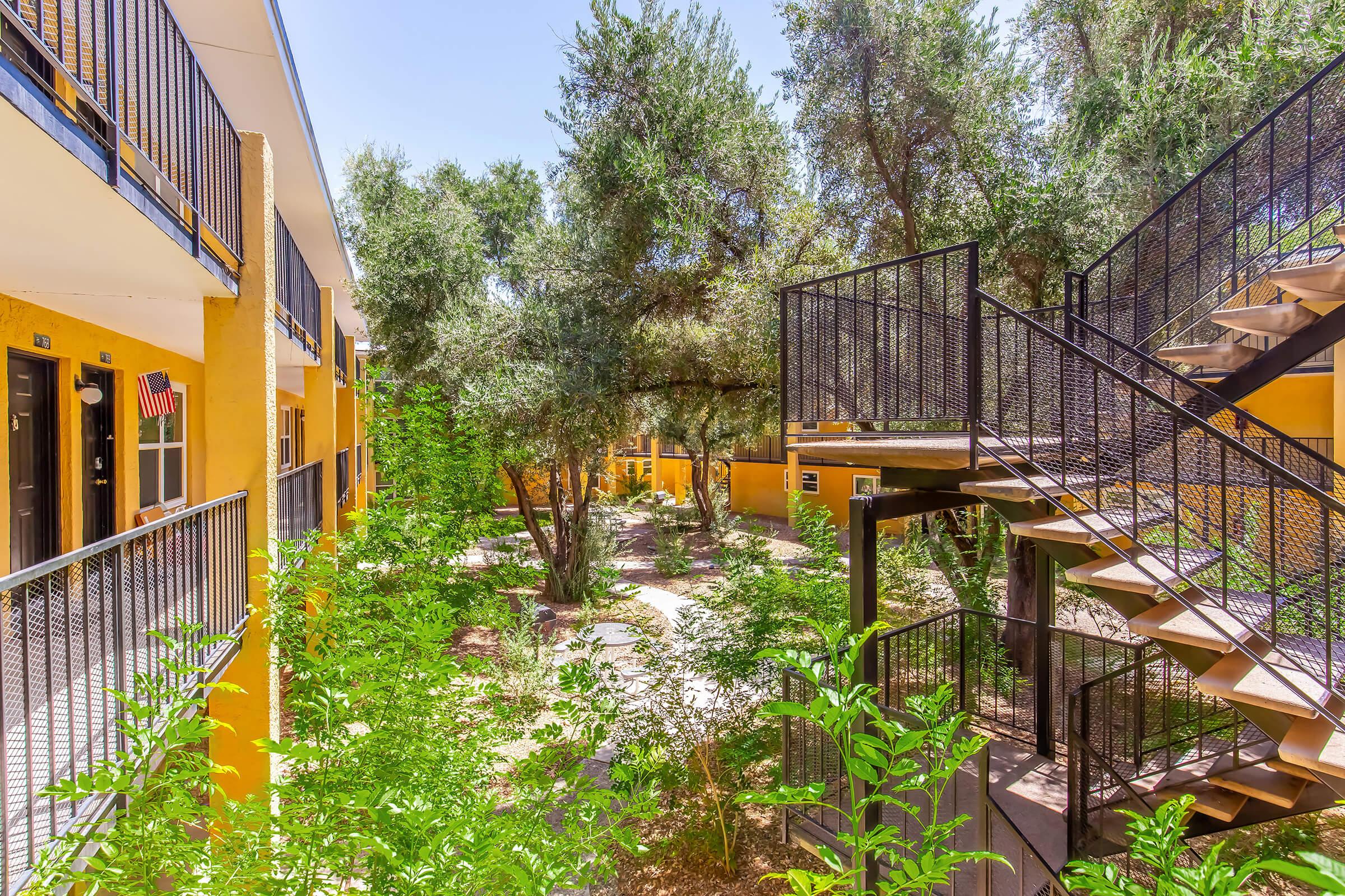A sunny courtyard surrounded by vibrant yellow buildings, featuring lush greenery, trees, and a metal staircase. The scene evokes a calm, inviting atmosphere, highlighted by blue skies and natural light filtering through the foliage.