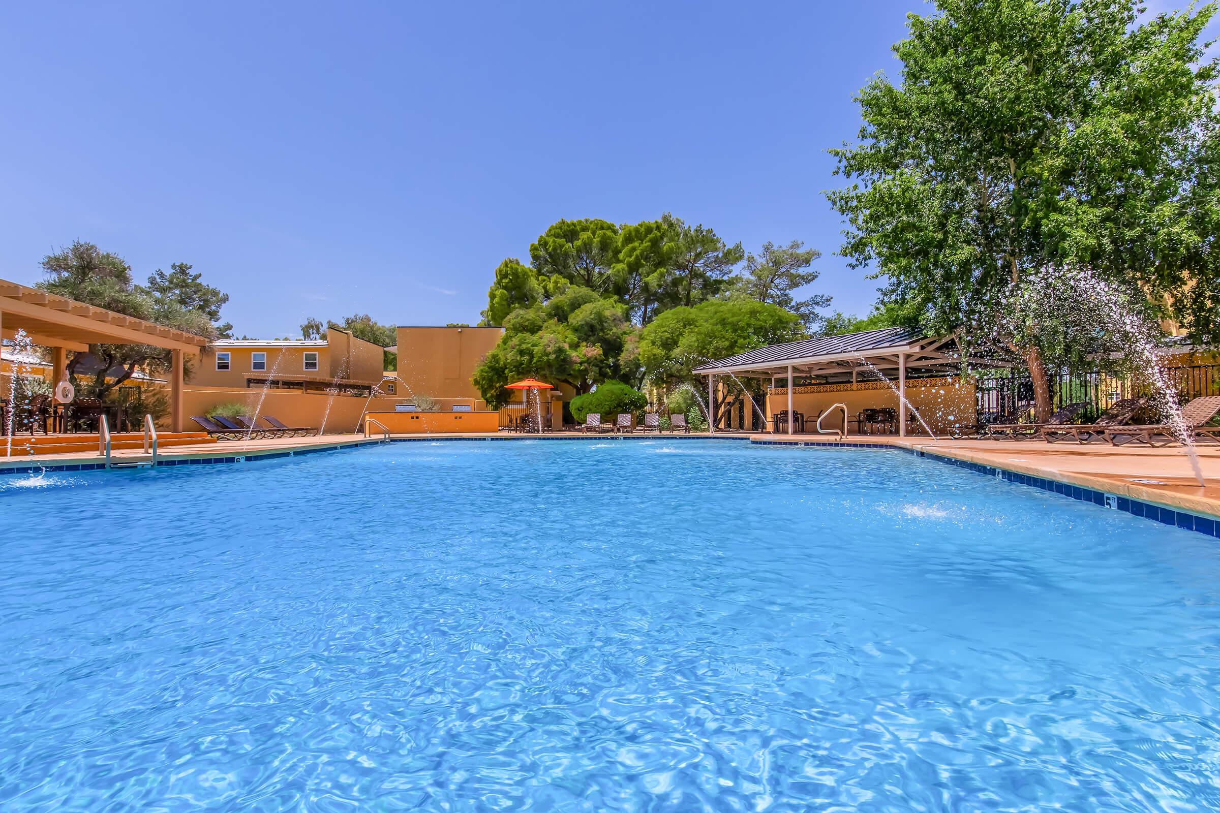 A clear blue swimming pool with a shaded area featuring lounge chairs, surrounded by lush greenery and bright orange umbrellas. The sky is sunny with a few scattered clouds, and the pool area is designed for relaxation and enjoyment.
