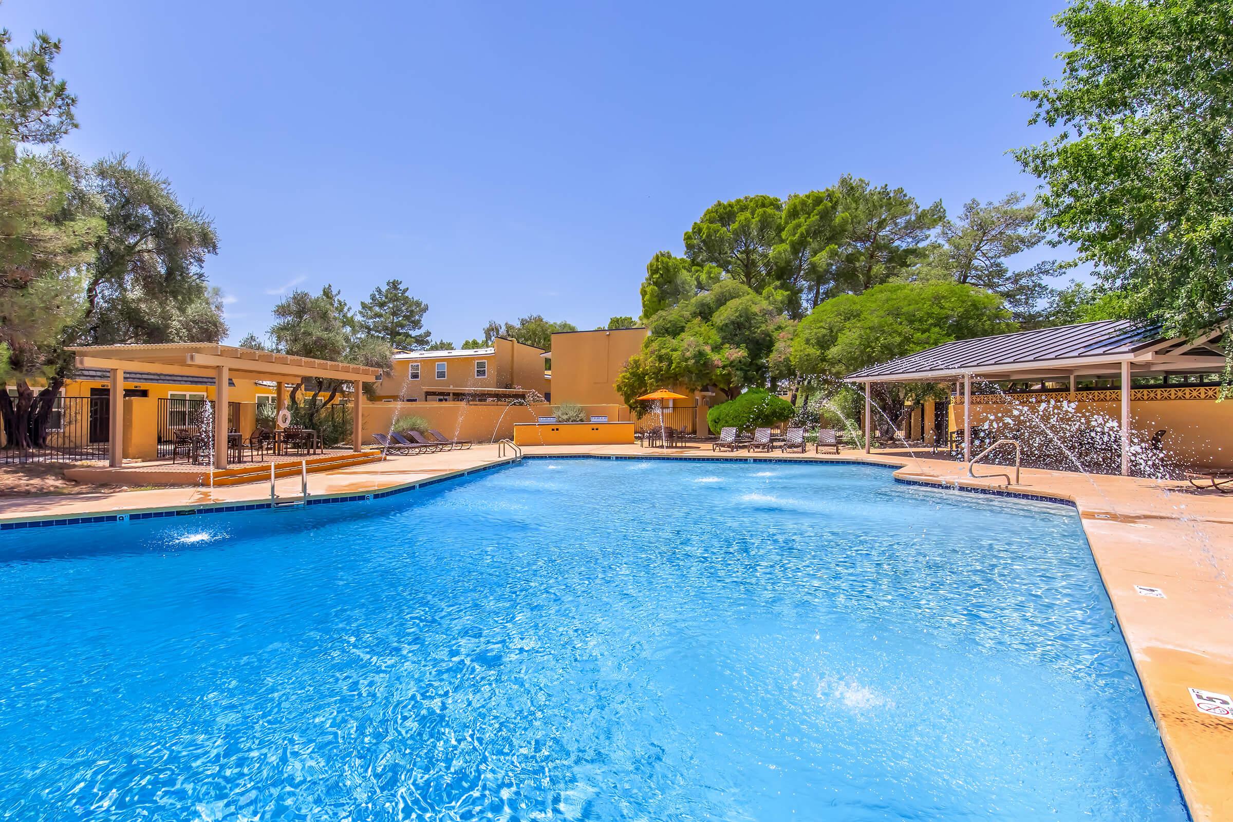 Swimming pool surrounded by lush greenery and orange-colored buildings. Sun loungers can be seen around the pool, with umbrellas providing shade. The clear blue water is highlighted by sunlight, and a splash feature adds a playful element to the scene. Bright, clear sky enhances the overall cheerful atmosphere.