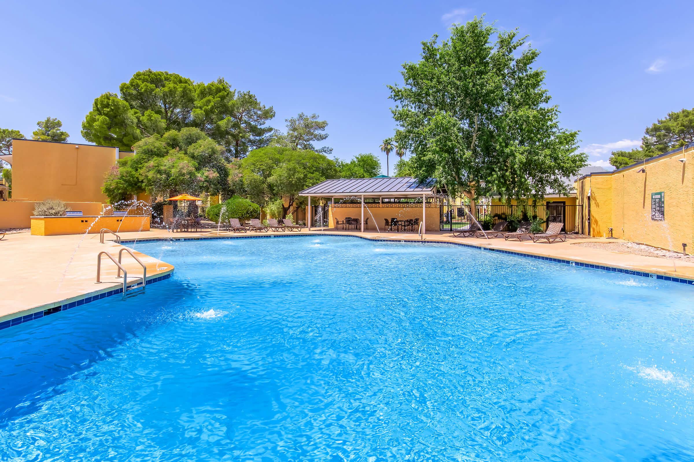 Outdoor swimming pool with clear blue water, surrounded by green trees and lounge chairs. A shaded gazebo is visible in the background, and the sky is bright and sunny with few clouds. The area appears well-maintained and inviting for leisure activities.