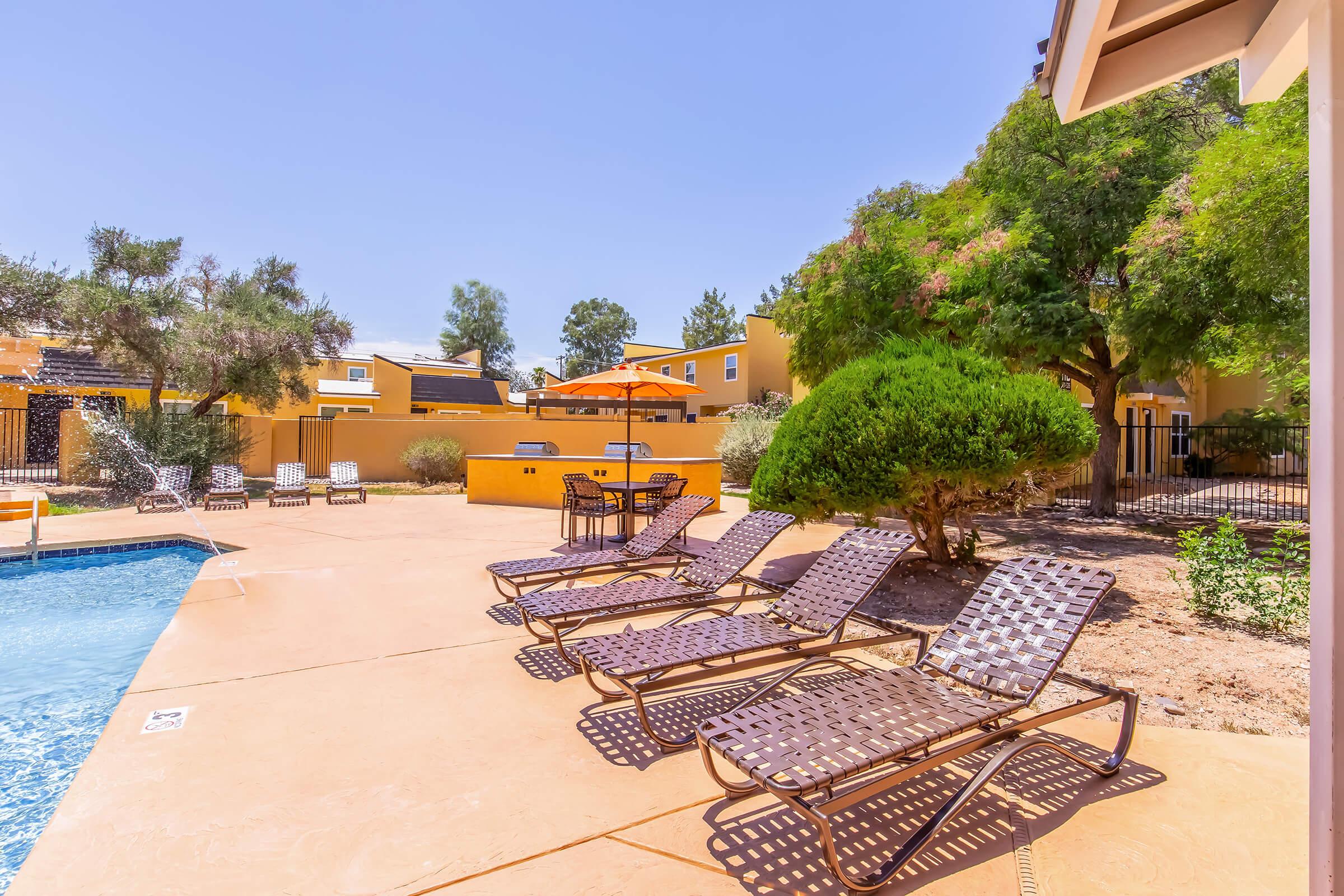A sunny outdoor swimming pool area featuring a clear blue water pool, several lounge chairs, and an orange umbrella. Surrounding the pool are landscaped areas with shrubs and trees, and yellow-finished buildings in the background. The scene is bright and inviting, perfect for relaxation.