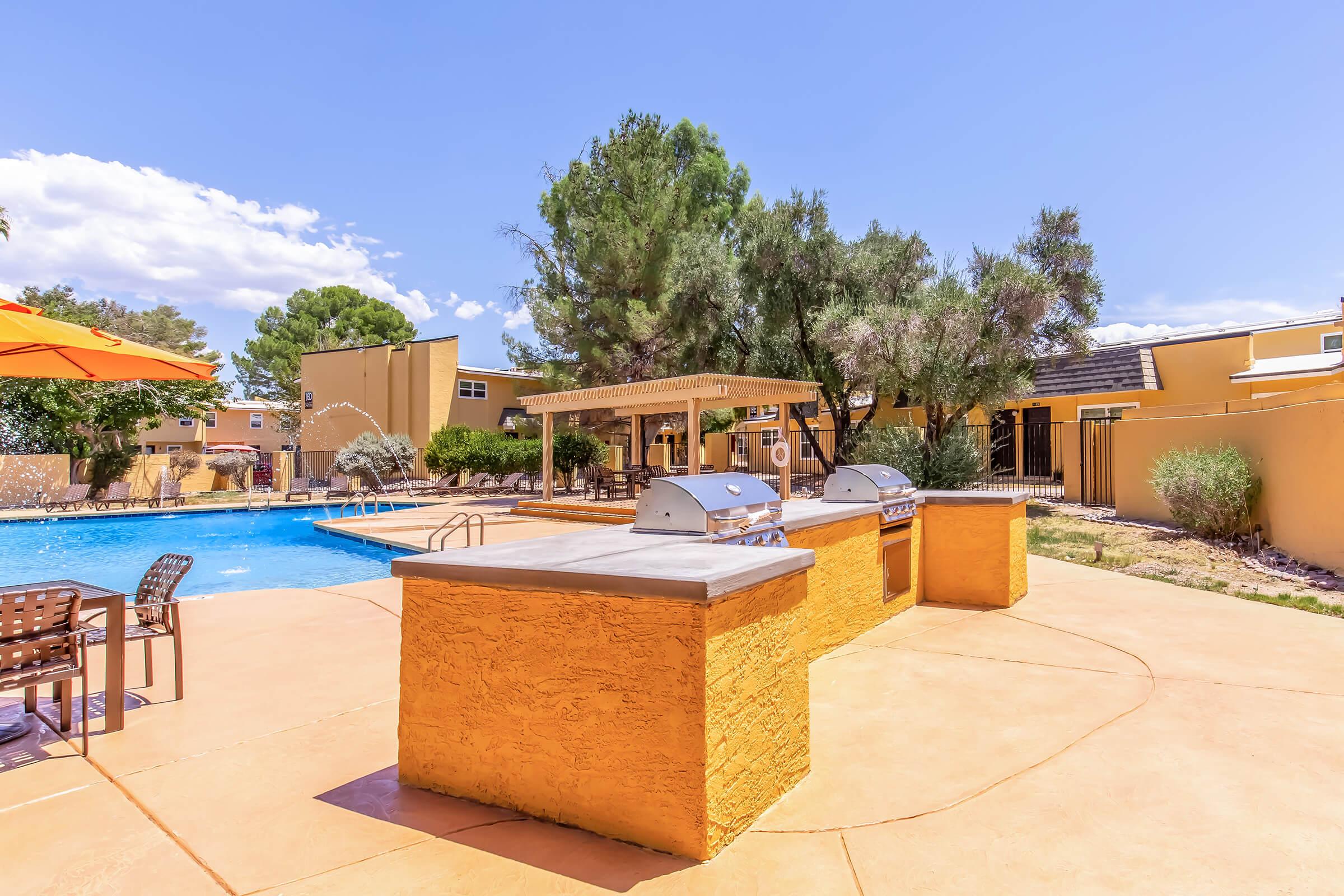Outdoor pool area featuring a swimming pool with a diving board, surrounded by lounge chairs and trees. A covered grilling station with two barbecue grills is placed on a textured patio. Bright blue sky with a few clouds above and a well-maintained property in the background.