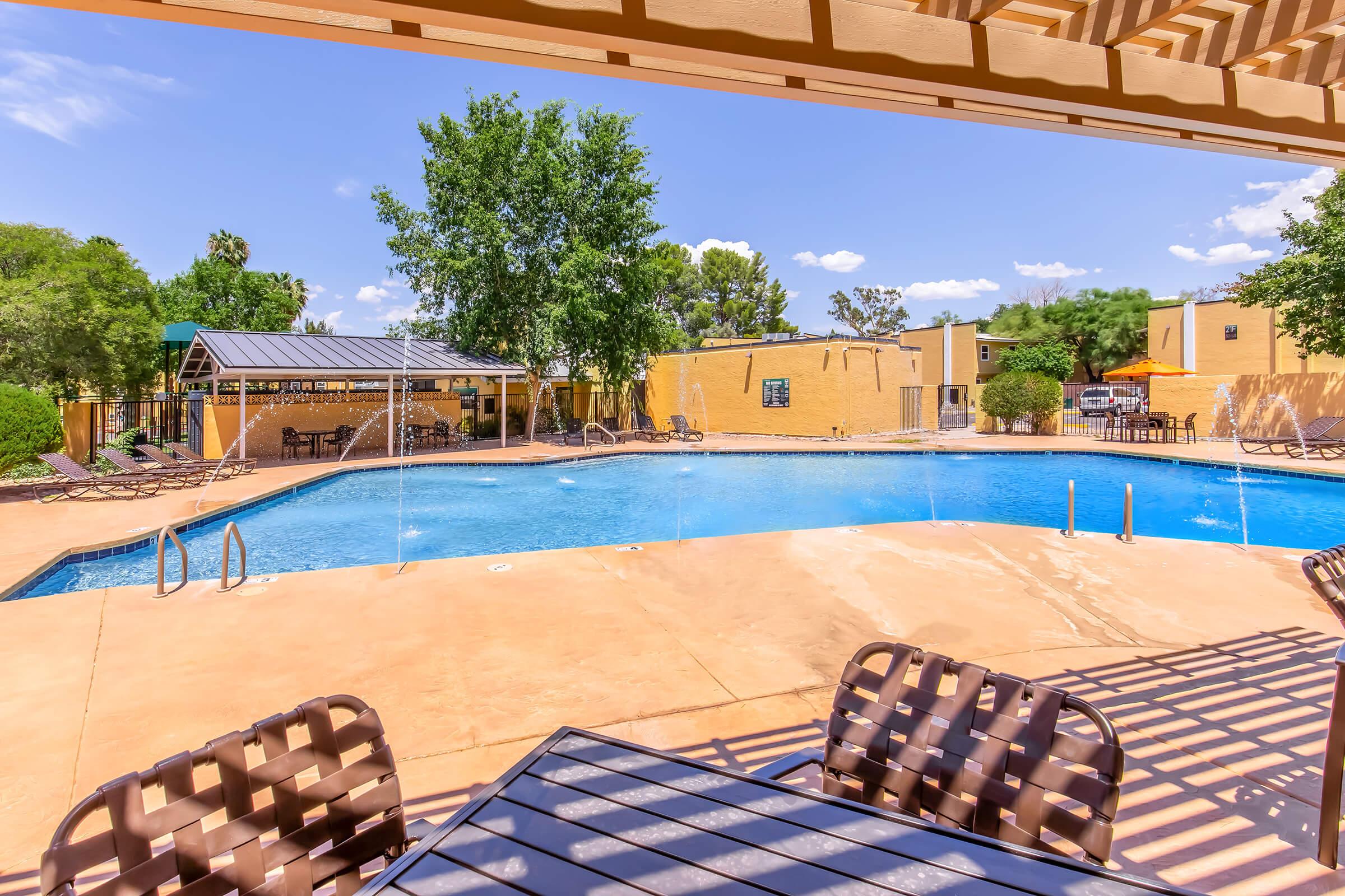 A sunny pool area featuring a sparkling blue pool with water fountains. Surrounding the pool are lounge chairs and shaded seating under a pergola. Lush greenery and a nearby gazebo enhance the relaxing atmosphere, while clear blue skies add to the inviting feel of the space.