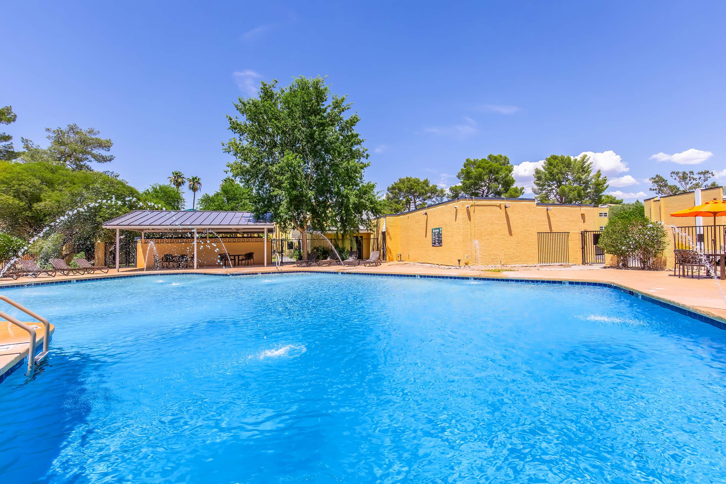 A bright and inviting swimming pool surrounded by trees and landscaped areas. Sunlight reflects off the clear blue water. In the background, there are yellow buildings and a shaded seating area with umbrellas. The atmosphere is serene and perfect for relaxation.