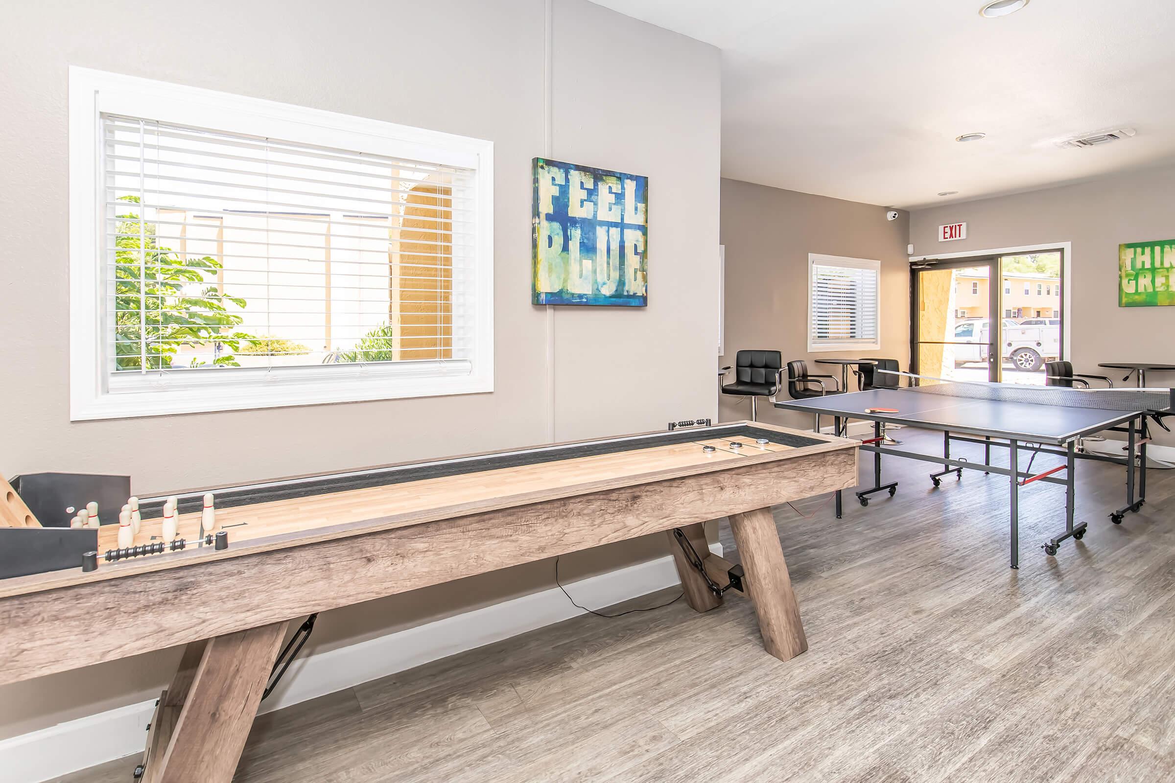 A spacious room featuring a shuffleboard table and a ping pong table, with natural light coming through a window. The walls display colorful artwork, including the phrases "FEEL BLUE" and "THINK GREEN." There are chairs and a doorway leading outside, creating a welcoming atmosphere.