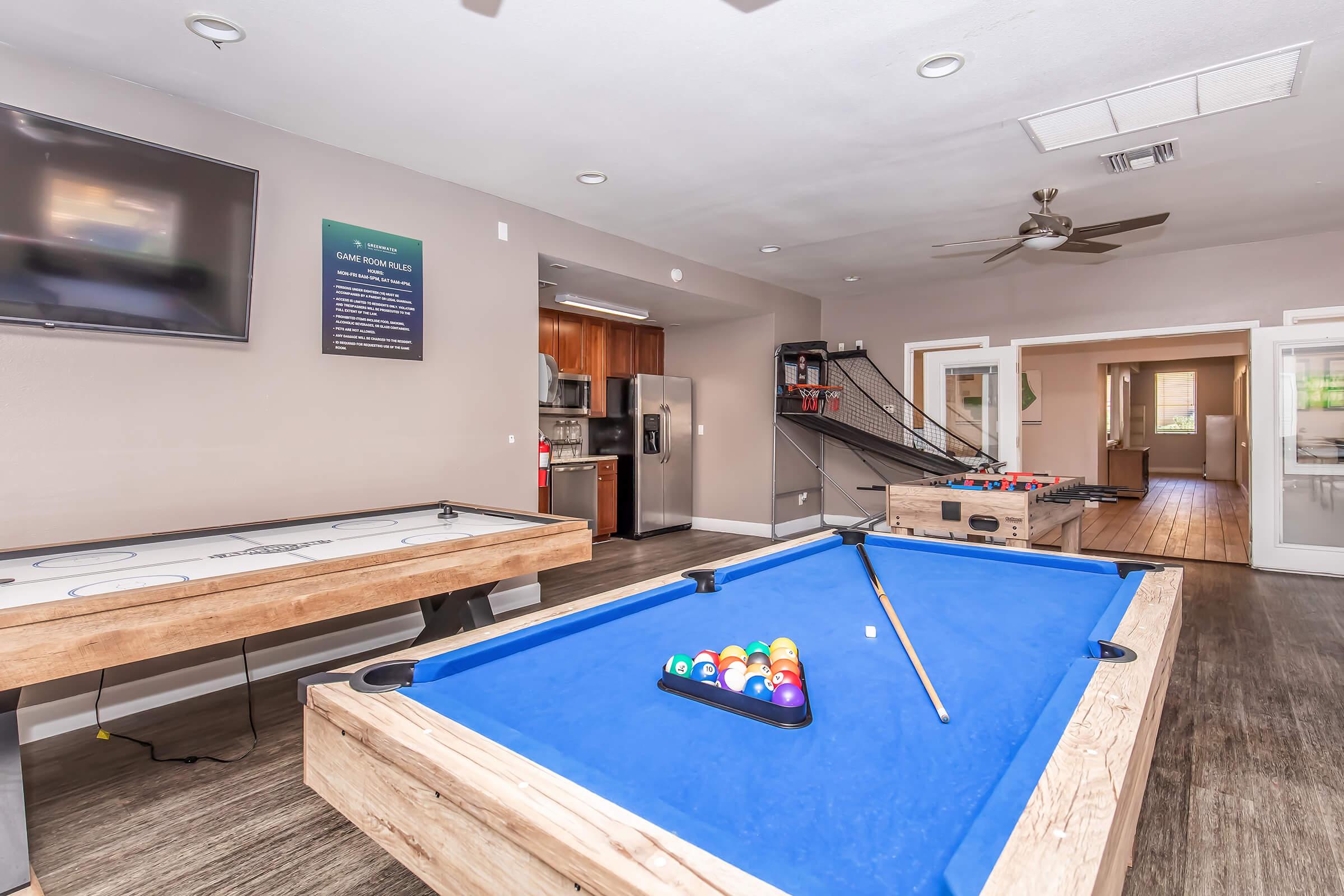 Interior of a recreational room featuring a blue billiards table with balls arranged, an air hockey table, a large TV mounted on the wall, and modern kitchen appliances visible in the background. The space is well-lit and includes wooden flooring and a staircase leading to another area.