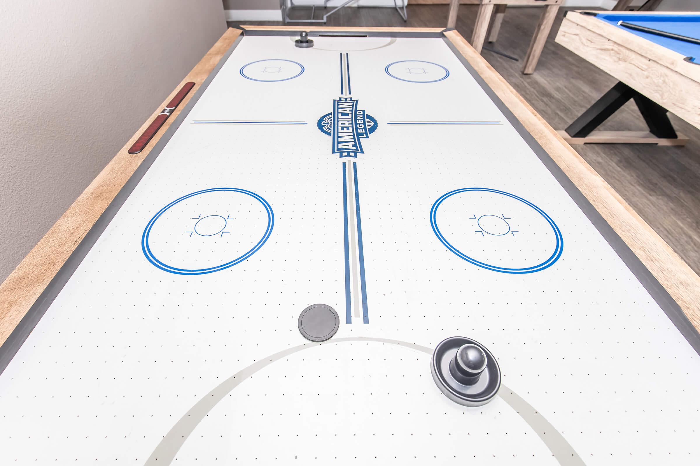 Air hockey table viewed from above, featuring a smooth white playing surface with blue circle markings, two puck goals, and a black puck and striker in position ready for play. The surrounding area includes a hint of wood texture from the table's edge.