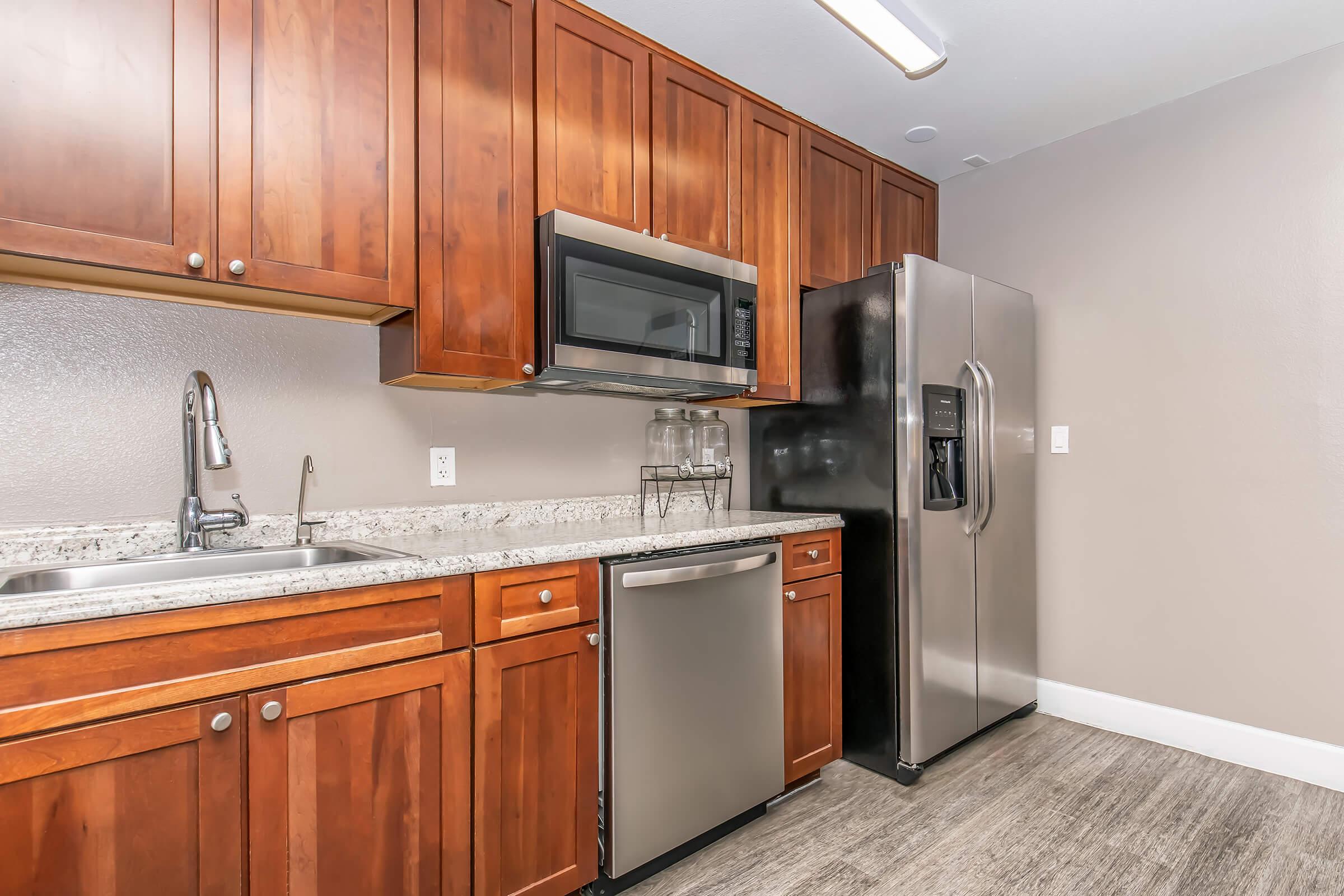 Modern kitchen with wooden cabinets, stainless steel appliances including a microwave, refrigerator, and dishwasher. The granite countertop features a sink with a faucet, and the space has neutral-colored walls and lighting. The flooring is a dark, textured design.