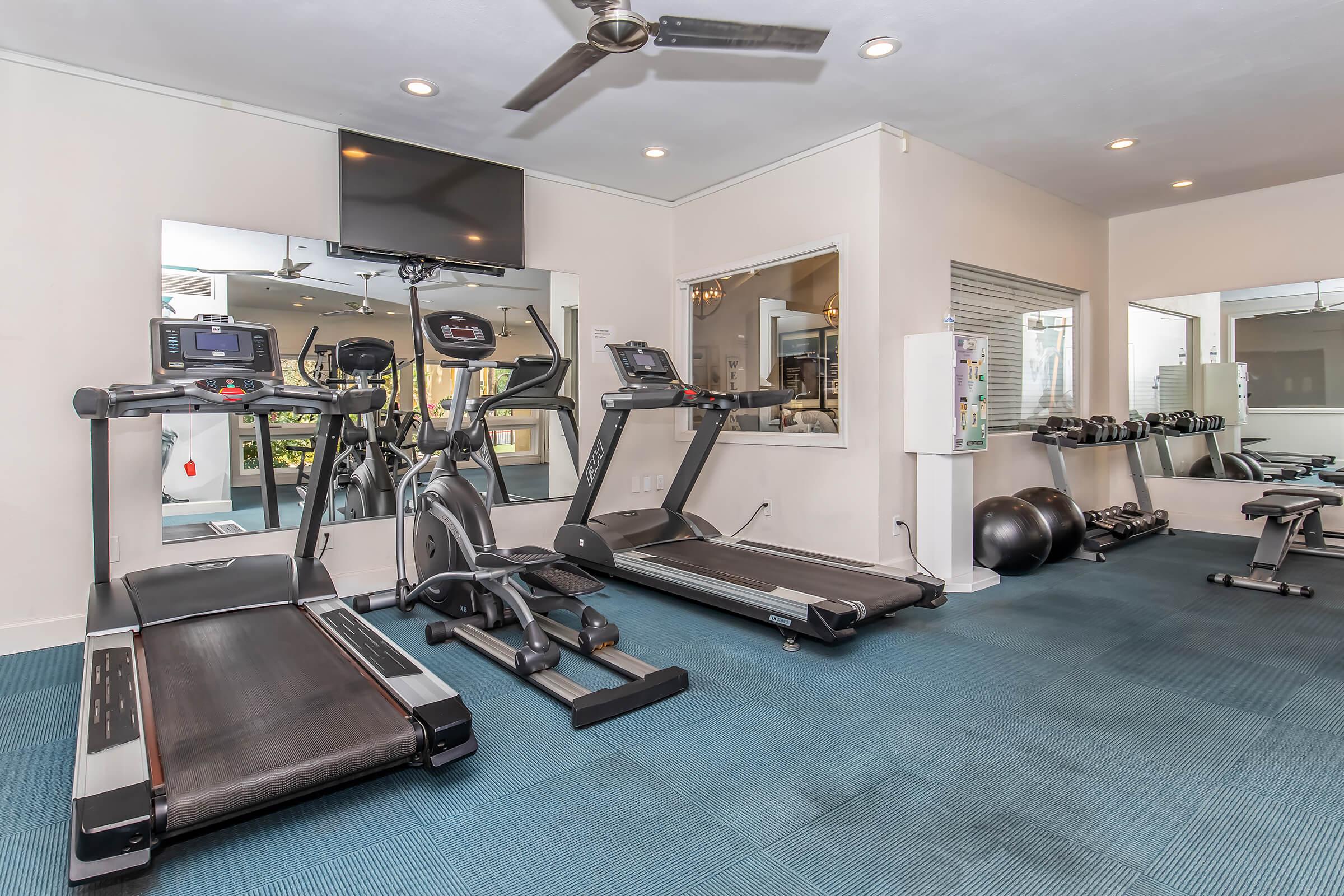 A modern gym interior featuring various exercise equipment including treadmills, an elliptical machine, free weights, and a weight bench, with large mirrors and a ceiling fan. The flooring is a blue carpet, and there's natural light coming in from windows.