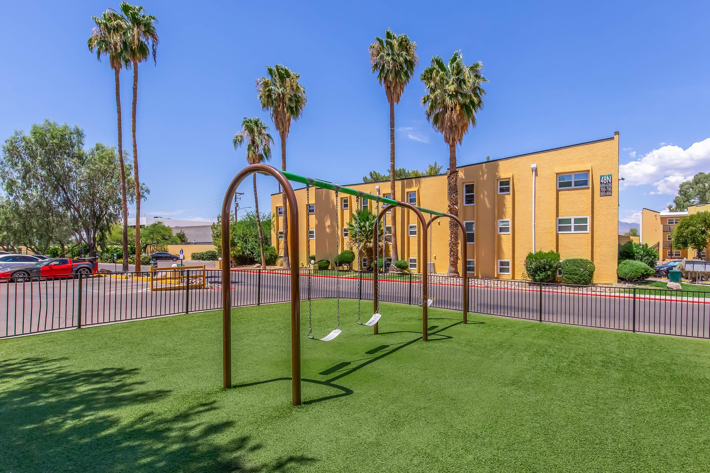 A playground featuring a set of swings on a green turf surface, surrounded by palm trees and colorful apartment buildings in the background under a clear blue sky.