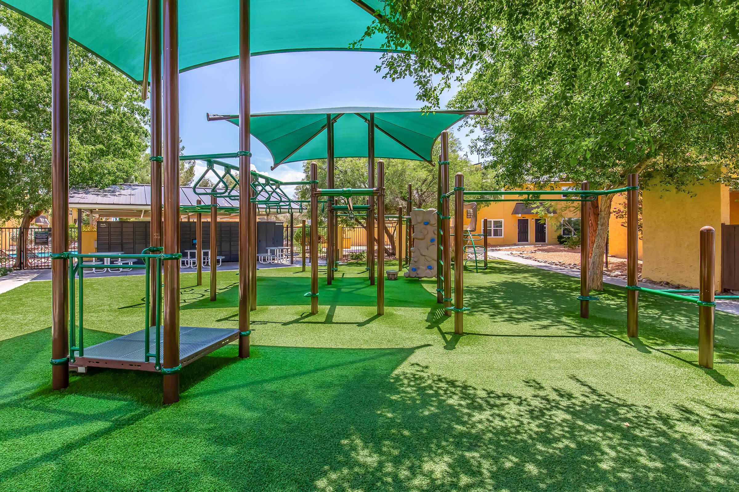 A vibrant playground featuring green artificial grass, modern climbing structures, and shade canopies. The area is surrounded by trees and a colorful building in the background, providing a welcoming environment for children to play and explore.