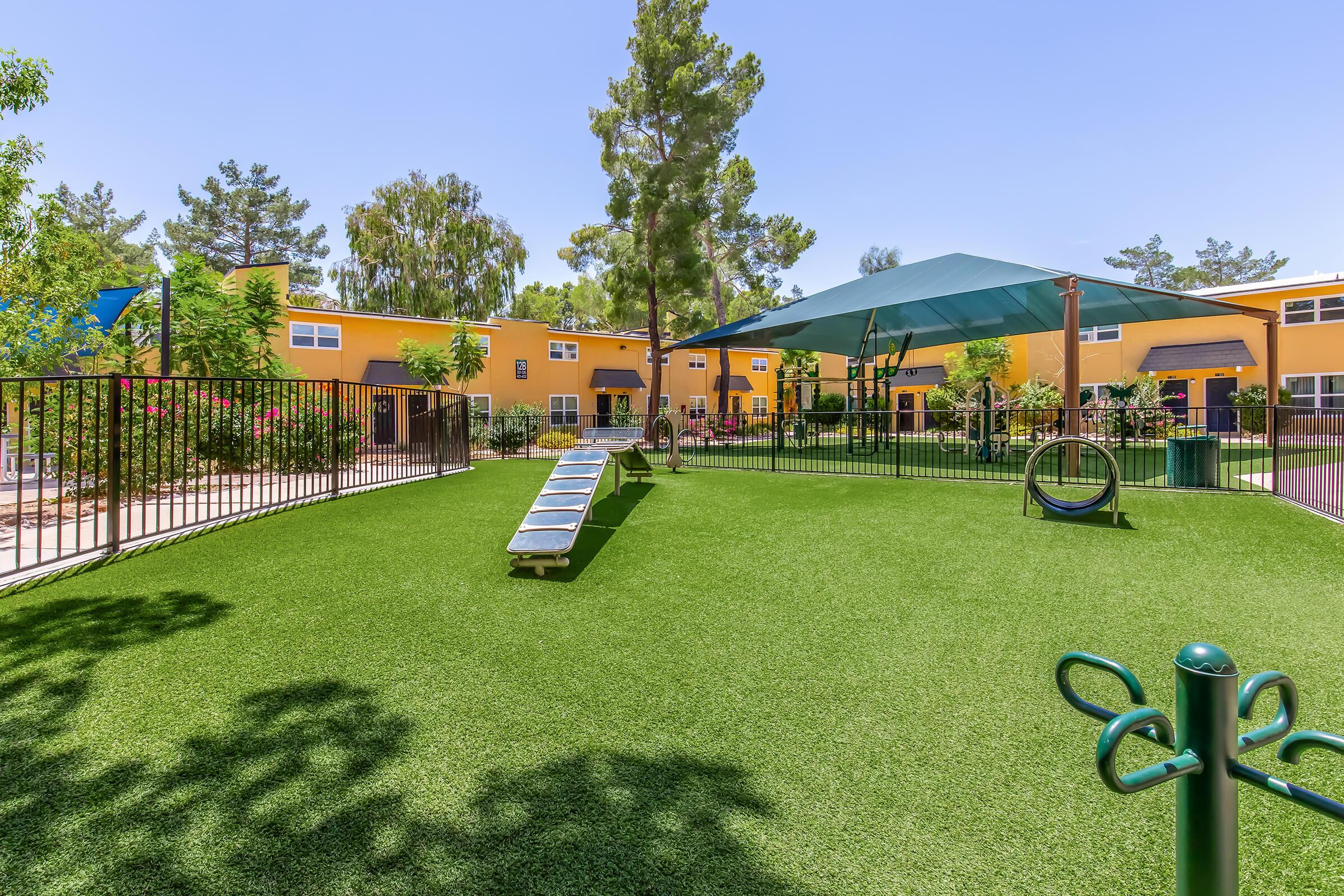 A vibrant playground area featuring green artificial grass. There is a set of lounge chairs, a shaded play structure, and various plants and trees surrounding colorful buildings in the background. The scene is bright and inviting, ideal for outdoor activities and relaxation.