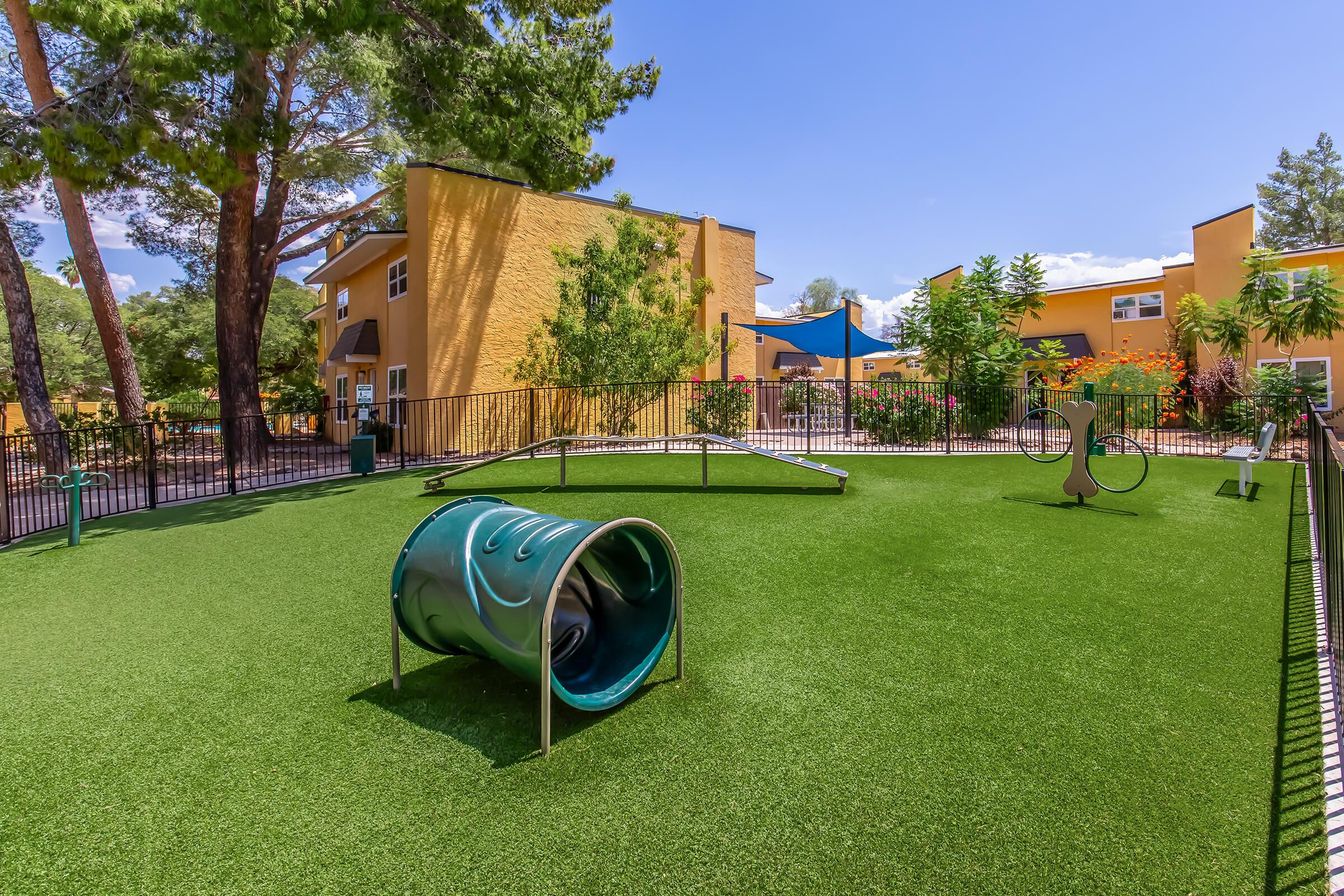A well-maintained dog park featuring artificial turf, a green cylindrical tunnel, an agility ramp, and a water station. The park is enclosed by a black fence, surrounded by trees and colorful flowers, with several attractive buildings in the background under a clear blue sky.
