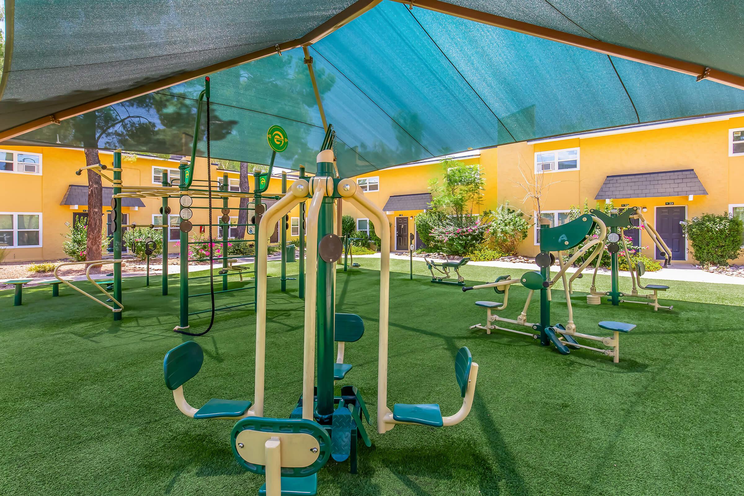 Outdoor fitness area with various exercise equipment under a shaded canopy, surrounded by green turf and colorful buildings in the background. The space is designed for physical activity and features machines for strength training and cardio.