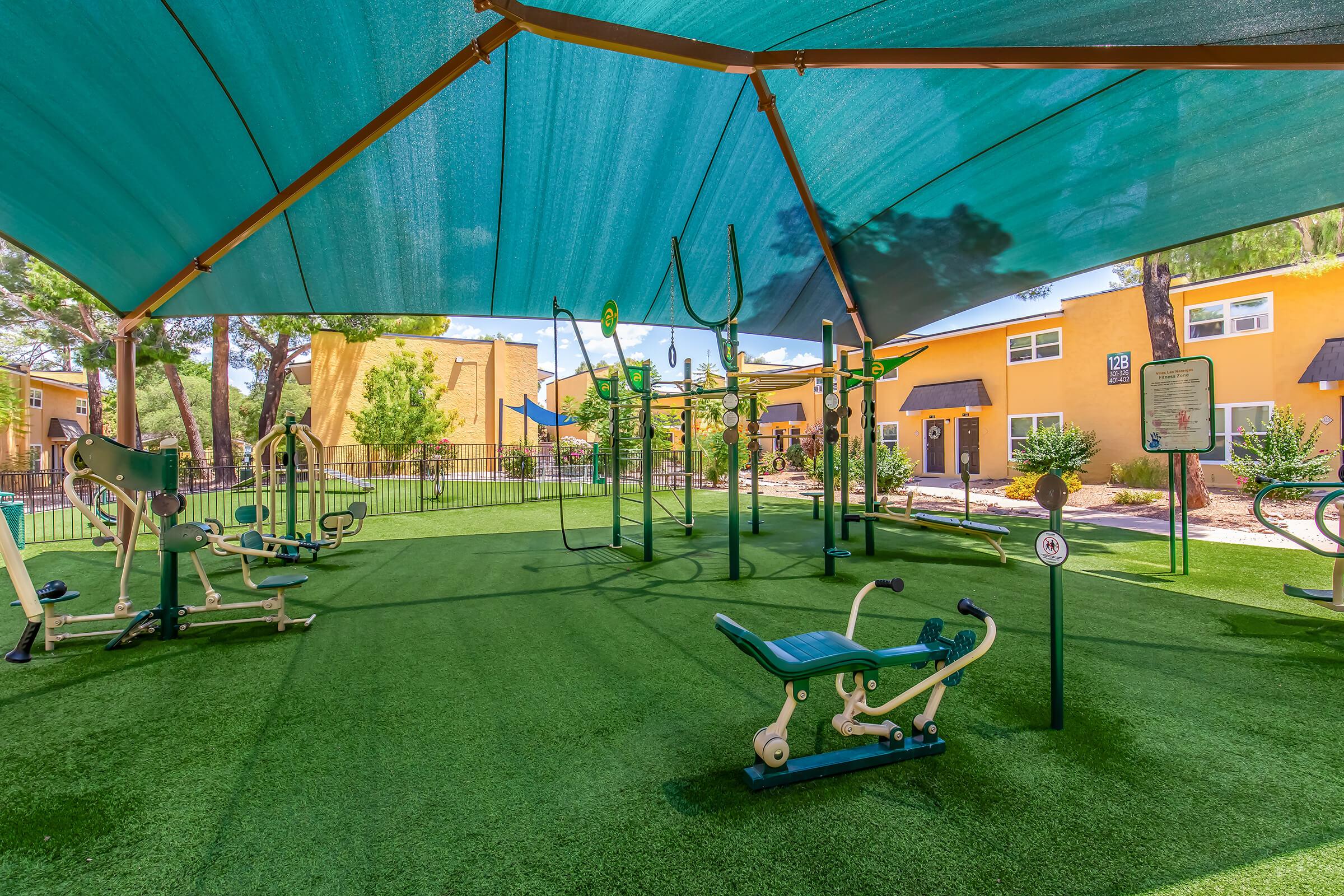 Outdoor fitness area featuring various exercise equipment under a large shaded canopy. The area has artificial turf and is surrounded by yellow buildings and a fence, creating a welcoming space for outdoor workouts.