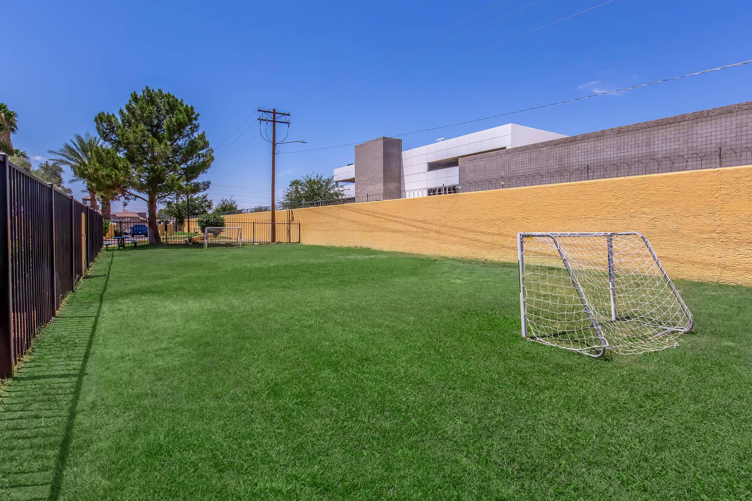 A grassy soccer field enclosed by a fence, featuring a small goalpost. In the background, there are trees and a building partially visible, under a clear blue sky. The scene conveys a calm, outdoor sports environment.