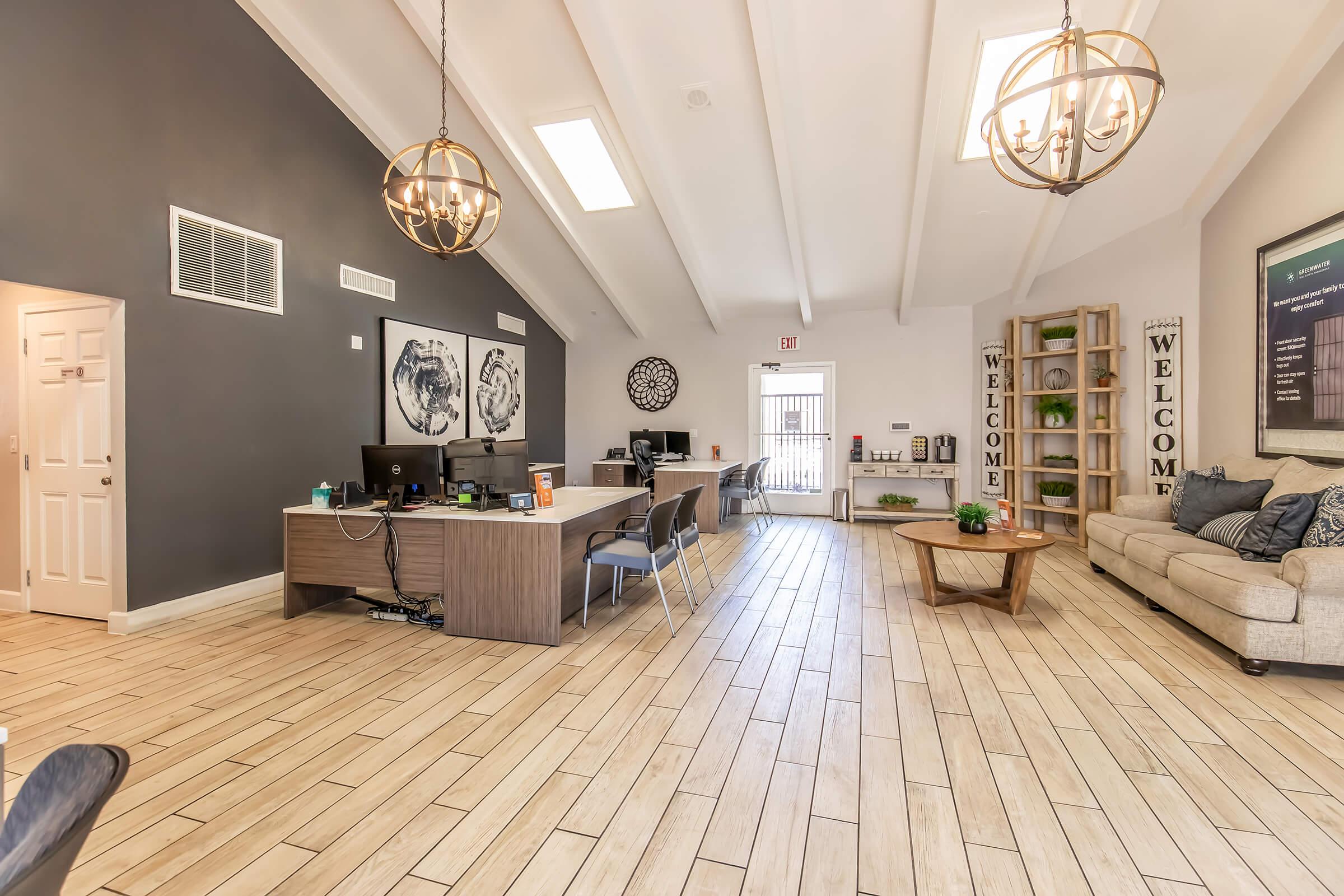 A spacious modern reception area featuring a large wooden desk with computers, comfortable seating, and decorative elements like potted plants and wall art. The ceiling is high with exposed beams, and there's natural light coming from skylights. A welcoming atmosphere is emphasized with the word "WELCOME" displayed on signs.