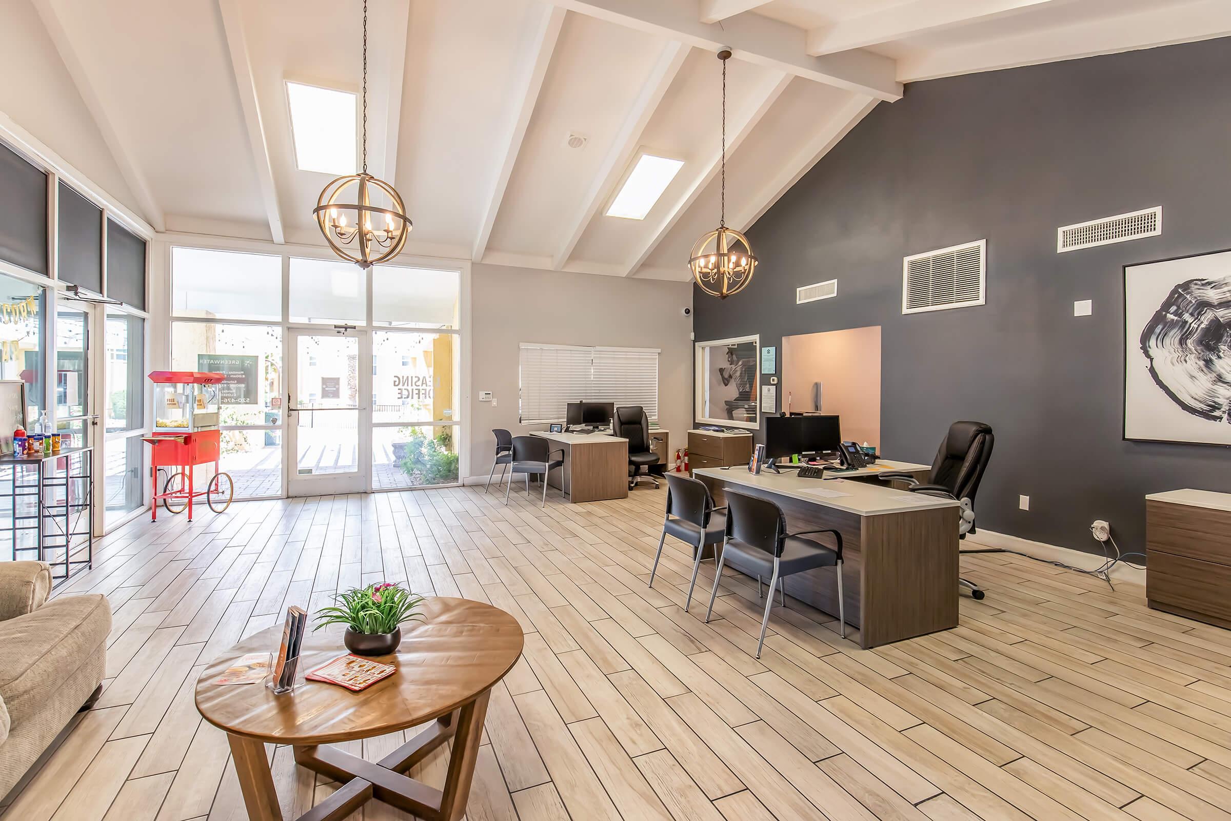 A modern office interior with a high, beamed ceiling. The space features light wood flooring, two desks with computers, and a cozy seating area. Large windows allow natural light in, and there is a decorative plant on the coffee table. A popcorn machine adds a playful touch to the environment.