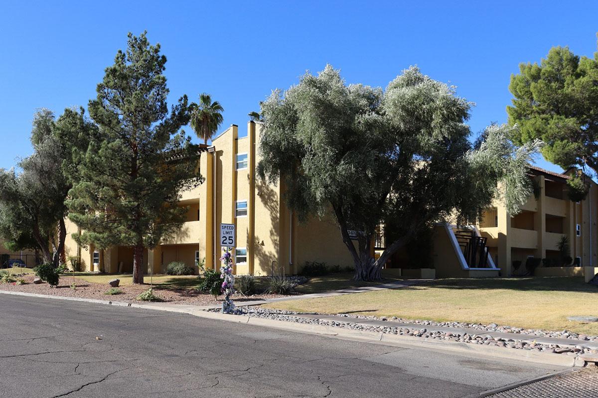 A view of a residential building surrounded by trees and greenery, located on a street with a 25 mph speed limit sign. The building features a yellow exterior and two levels, with shaded areas created by the nearby trees. Clear blue sky in the background.
