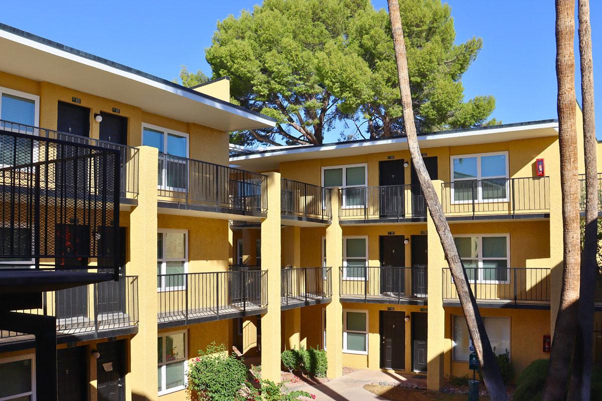 A view of a multi-story apartment complex featuring yellow exterior walls, black railings, and balconies. The scene is framed by tall pine trees, with clear blue skies in the background. Lush greenery and flower beds are visible in the courtyard area, creating a welcoming atmosphere.
