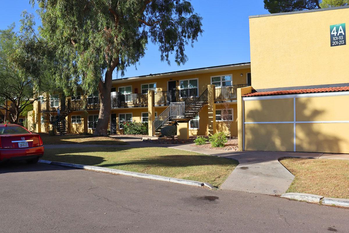 A view of a two-story apartment building with yellow exterior and balconies. A red car is parked on the street alongside the structure. Sunny weather with clear blue skies and green trees in the vicinity. The image also includes a sign labeled "4A" on the building.