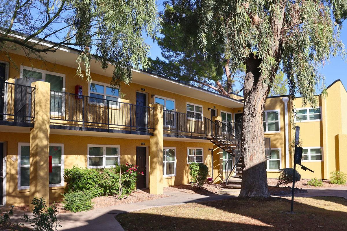 A view of a two-story apartment complex with yellow exterior walls. The building features balconies with black railings, surrounded by trees and shrubs. A staircase leads to the second floor, and there is a pathway leading through the landscaping. The scene is bright and well-maintained, reflecting a residential area.