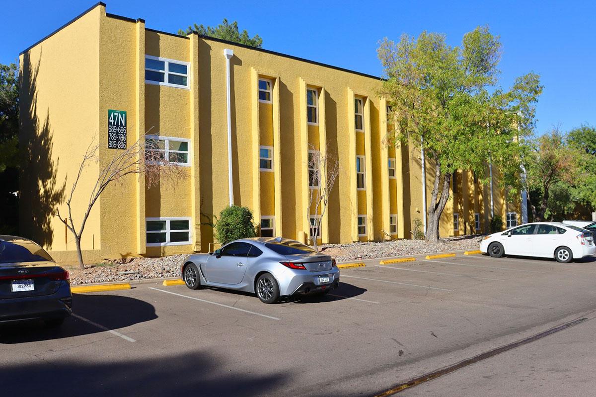 A modern yellow building with six vertical panels, surrounded by trees and parked cars. In the foreground, there are three vehicles: a silver sports car on the left, a white car on the right, and a dark-colored car nearby. The clear blue sky provides a vibrant backdrop.