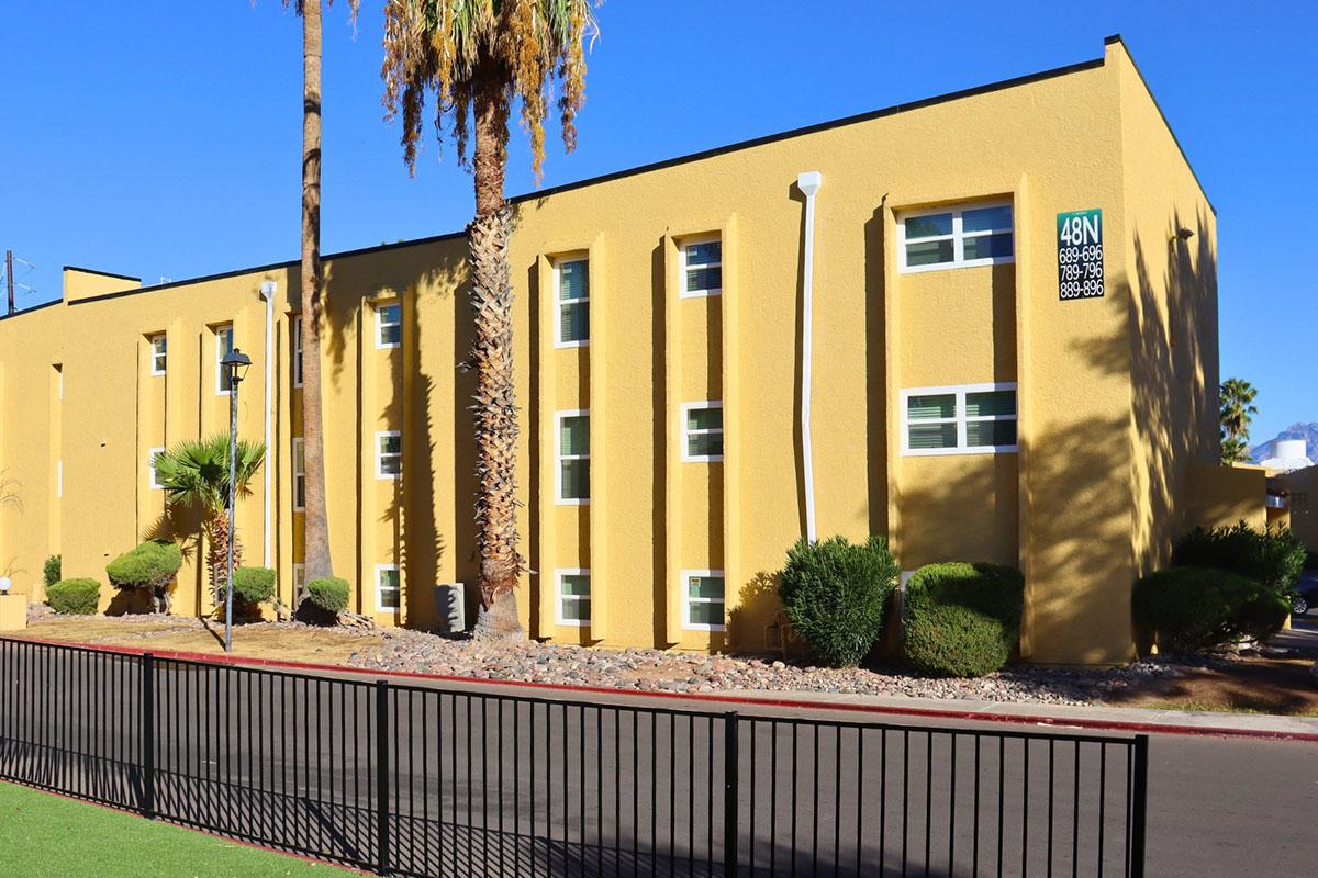 A yellow, two-story building with multiple windows surrounded by palm trees and desert landscaping. A green lawn is in the foreground, and a black fence runs along the road in front of the building. A street sign indicates "48N."