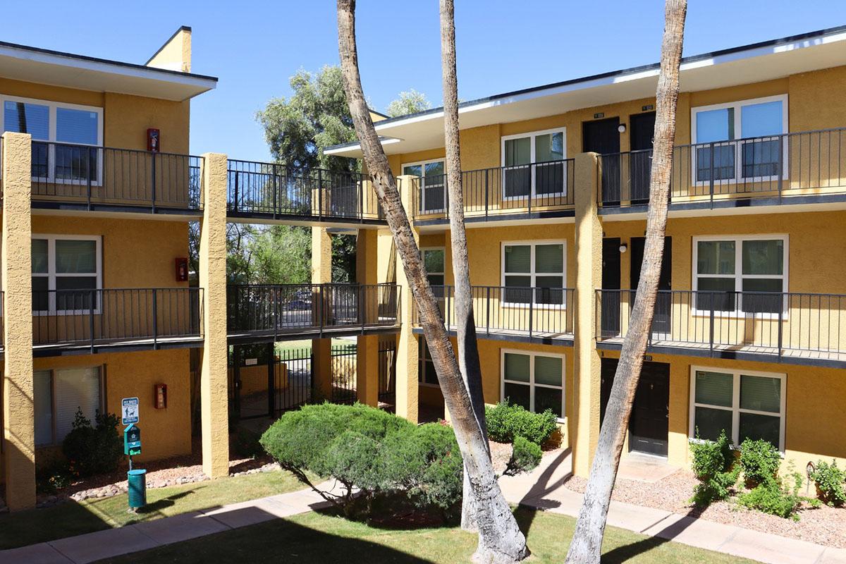View of a residential complex featuring two buildings with yellow exteriors, balconies, and green landscaping. The scene includes palm trees and a pathway, highlighting the outdoor area between the two structures. The sky is clear and blue, suggesting a warm climate.