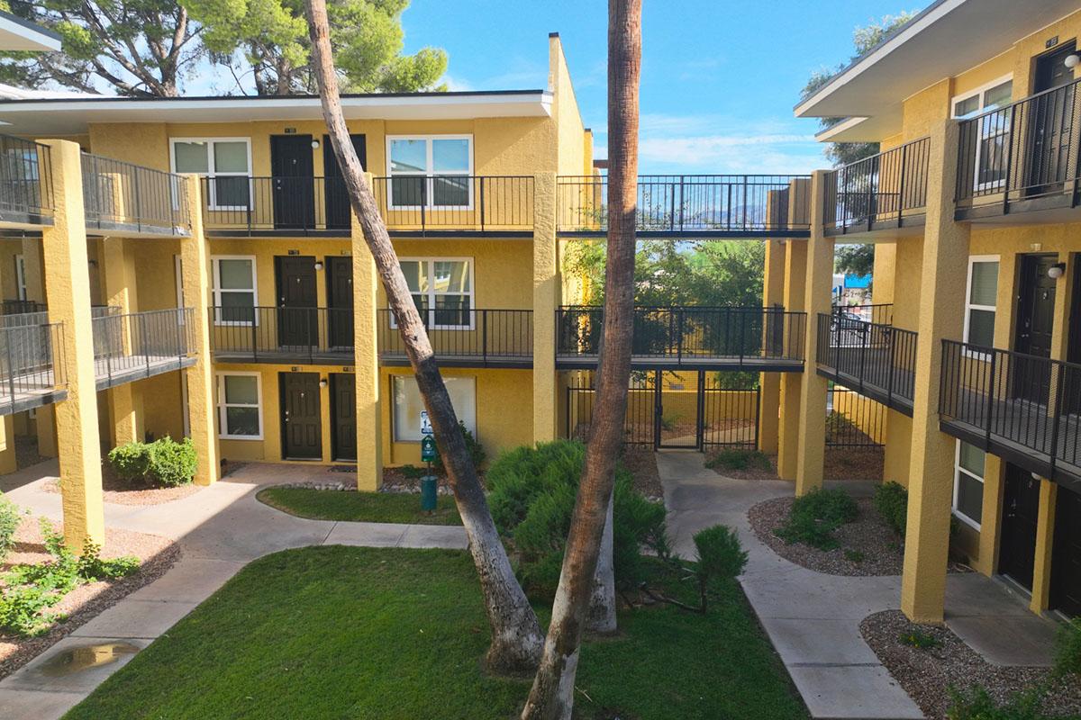 View of a yellow multi-story apartment complex surrounded by greenery. The scene shows a central courtyard with a paved path, trees, and balconies overlooking the area. Clear blue sky and bright sunlight enhance the inviting atmosphere of the location.