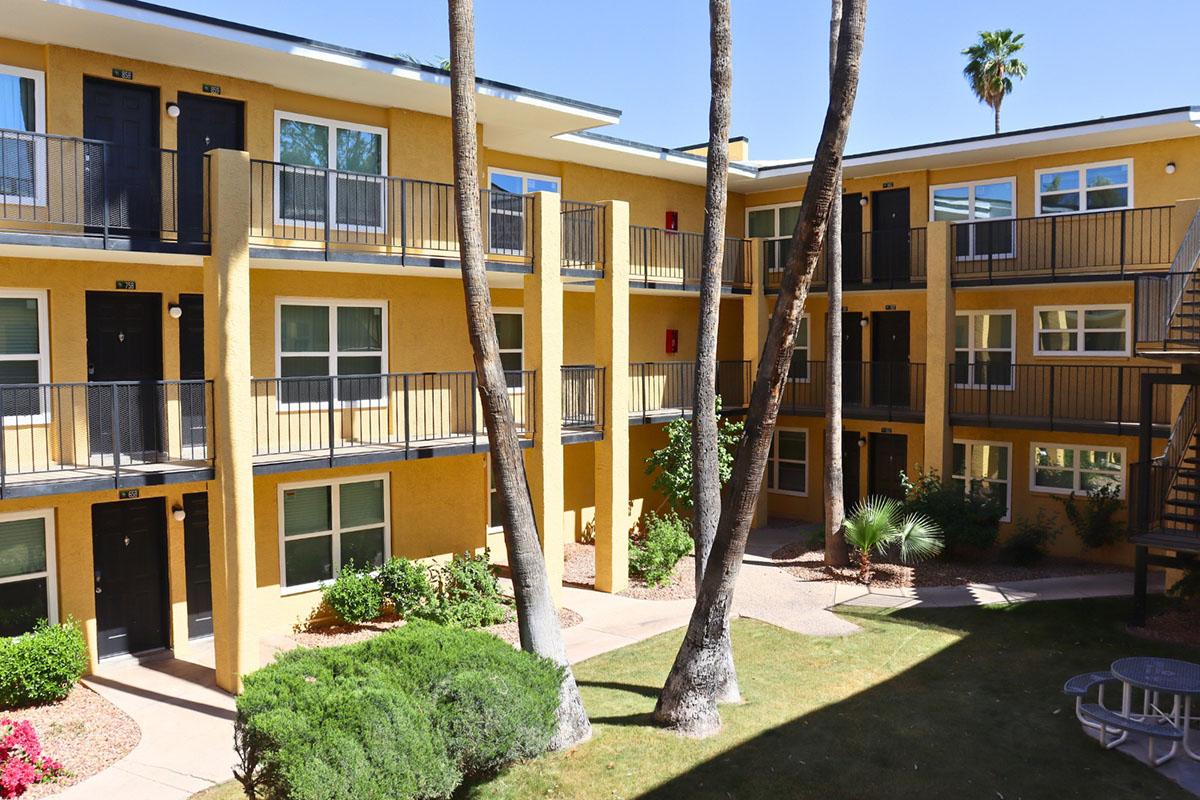View of a courtyard area in an apartment complex featuring multiple floors of yellow buildings. Balconies with black railings are visible, along with palm trees and landscaped greenery. A pathway leads through the garden space, creating a bright, sunny ambiance.