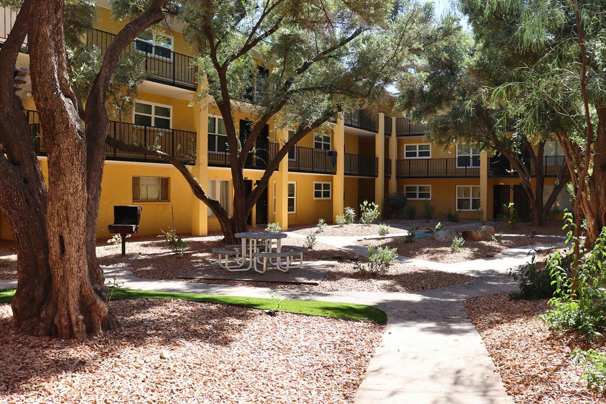 A landscaped courtyard of an apartment complex featuring several trees, a picnic table, and a pathway. The area is surrounded by yellow-painted buildings, with a mix of gravel and greenery along the ground.