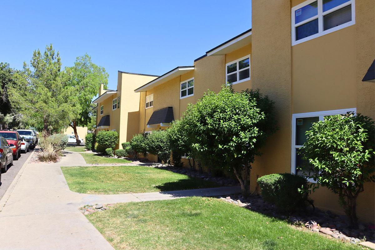 A row of yellow apartment buildings lined with green shrubs and trees, set against a clear blue sky. A pathway runs alongside the buildings, leading to grassy areas. Parked cars can be seen nearby, adding to the suburban atmosphere.