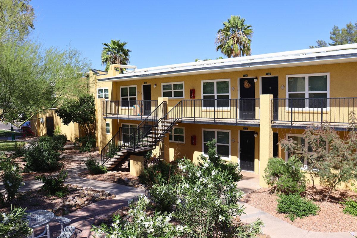 A sunny view of a multi-unit apartment complex featuring yellow buildings with black doors and railings. There is a staircase in the foreground, surrounded by well-kept landscaping including shrubs and palm trees. The sky is clear blue, indicating a warm day.