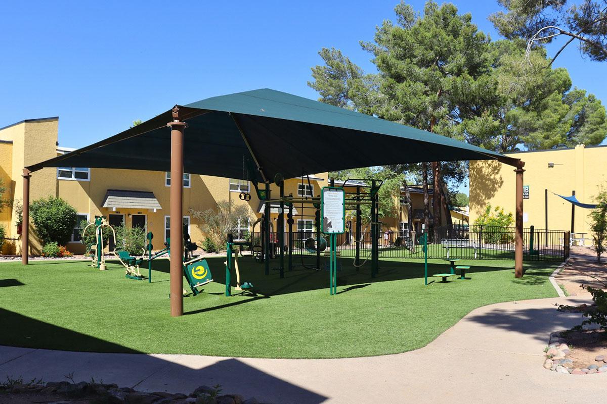 A shaded outdoor exercise area featuring various workout equipment surrounded by green grass and trees. The space is designed for fitness activities, with a protective canopy overhead and nearby residential buildings in a sunny environment.
