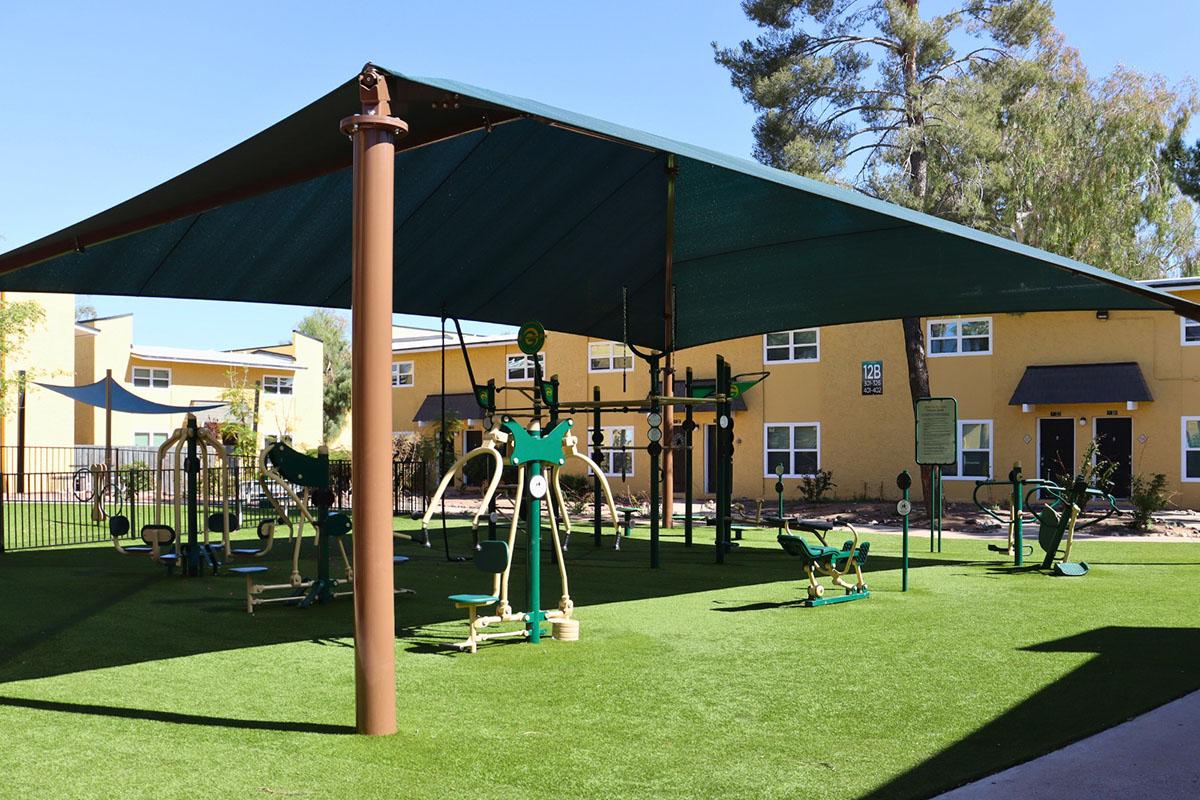 Outdoor fitness area with various exercise equipment under a large green shade structure. The space features artificial grass, surrounded by a fence and residential buildings in the background, providing a community-friendly atmosphere for exercise and recreation.