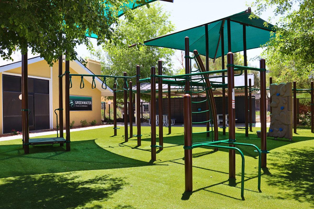 A colorful playground featuring climbing structures, slides, and swings, set on lush green artificial grass. The area is shaded by large canopies, with nearby trees providing additional shade. A wall for climbing is visible on one side, alongside a sign reading "Greenwater."