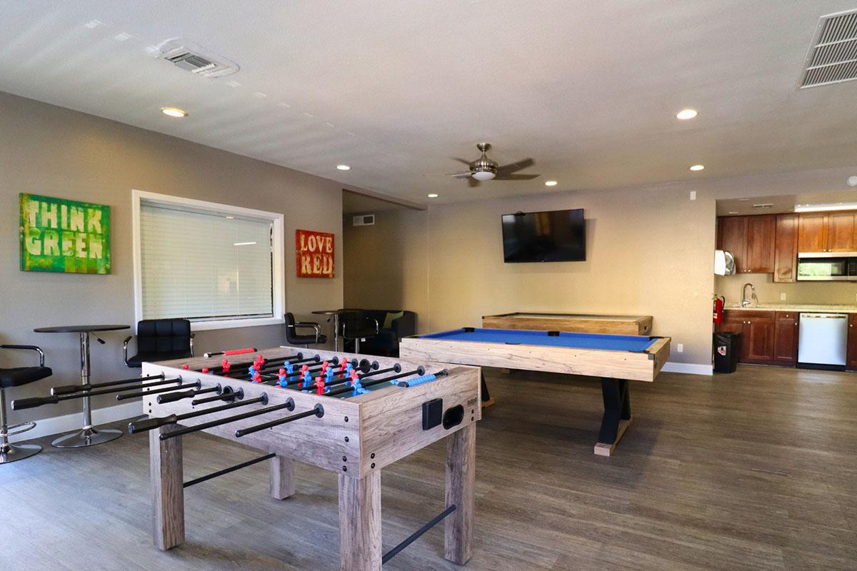 A spacious game room featuring a foosball table, a pool table, and a flat-screen TV. The walls are adorned with colorful motivational signs. There are modern bar stools and a sleek chair, with warm lighting and wood flooring creating a welcoming atmosphere.