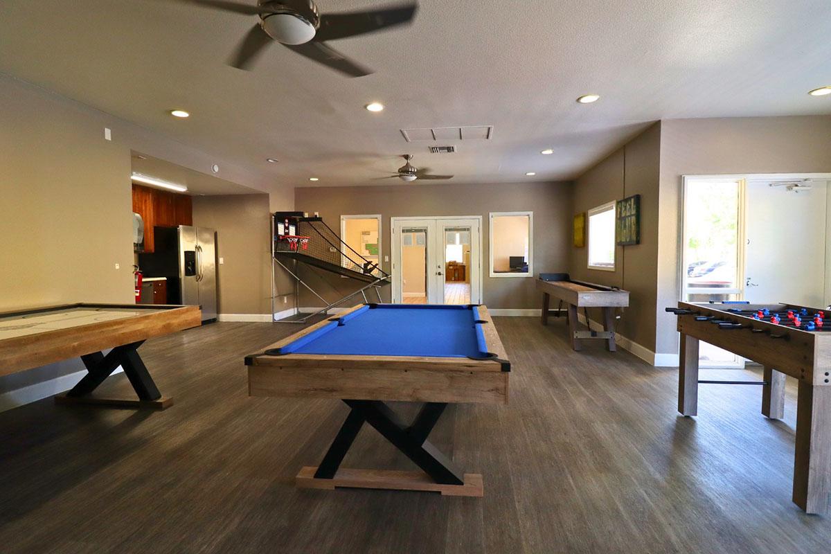 A spacious game room featuring a blue billiards table, a foosball table, and two wooden tables. The room has wooden flooring, beige walls, and ceiling fans. Natural light enters from a door on one side, and there is a small kitchenette visible in the background.