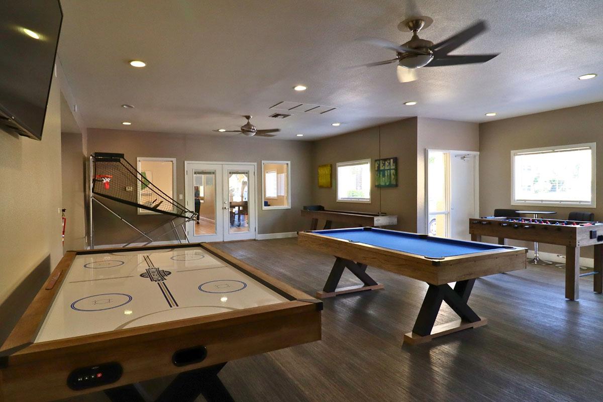 A recreational room featuring an air hockey table and a pool table made of wood. There is a foosball table in the corner, and a large television mounted on the wall. The space is well-lit with ceiling fans and has large windows allowing natural light. The walls are decorated with simple artwork.