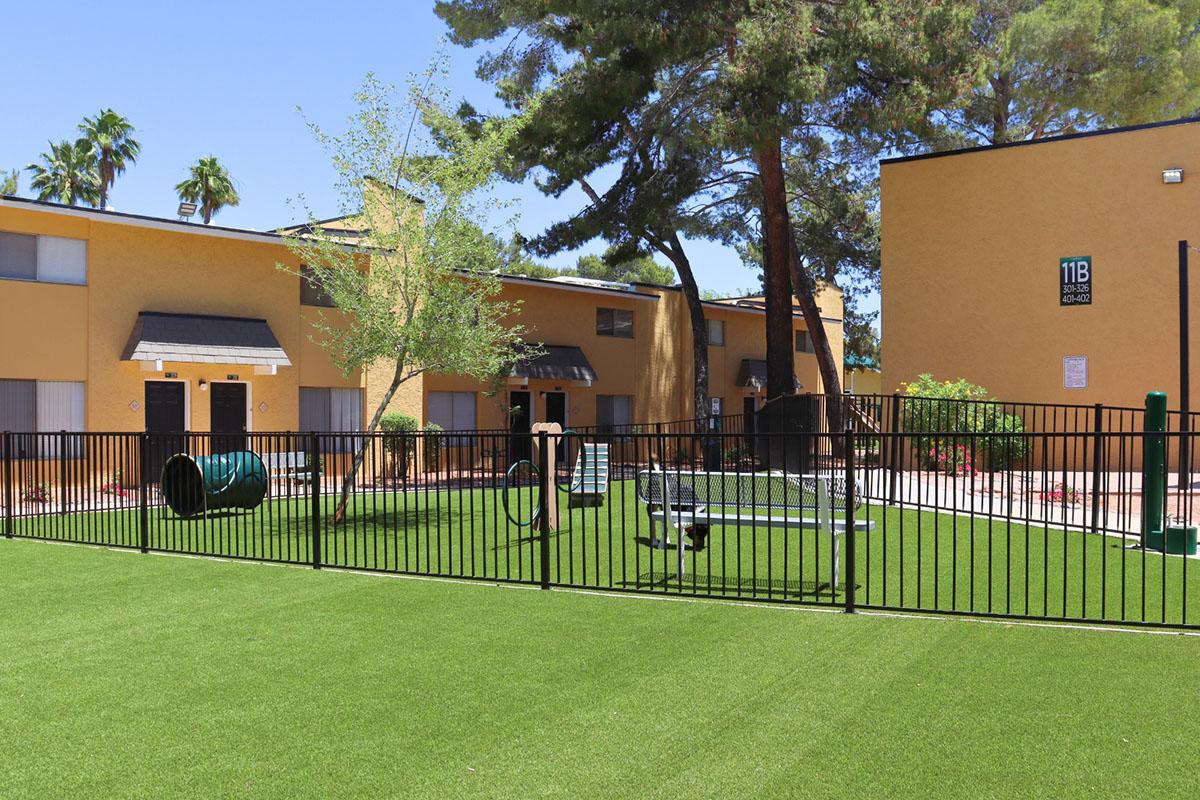 A well-maintained grassy area enclosed by a black fence, featuring a small playground with equipment. In the background, two yellow buildings are visible, surrounded by palm trees. The scene is bright and sunny, suggesting a pleasant outdoor space for residents or children to enjoy.